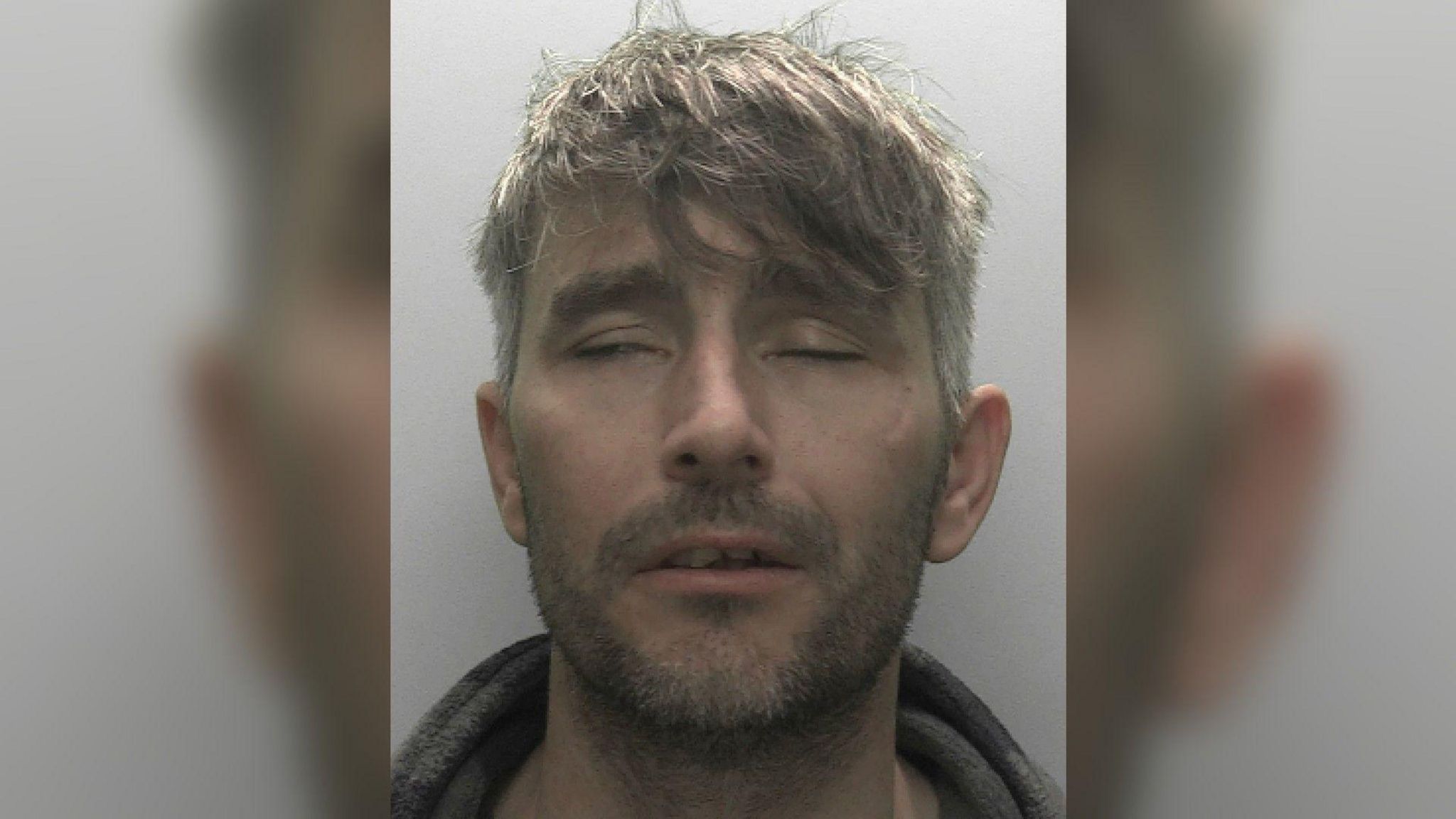 Mugshot of Jonathan Walsh, one eye closed the other slightly open, scruffy brown and grey hair, scruffy beard and mouth slightly open, grey background wearing a grey hoodie