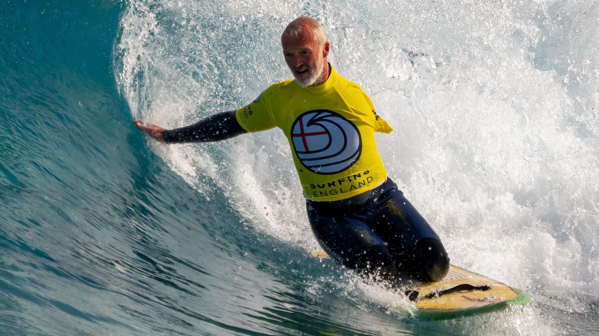 Mr Downes riding a wave that's curving over him on knees . It's a sunny day with the blue water sparkling as he trails his right hand through the water.