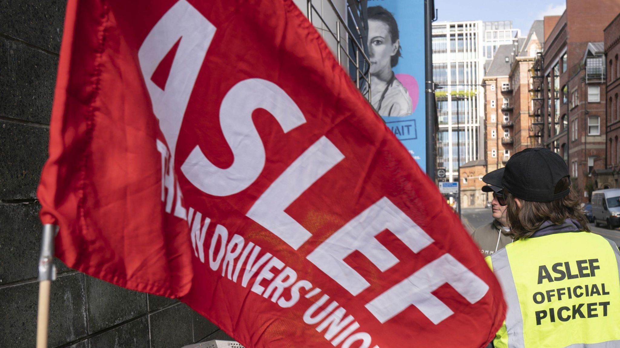 An Aslef flag, with a member of the union having his back to the camera, wearing a h-vis jacket reading "ASLEF OFFICIAL PICKET"