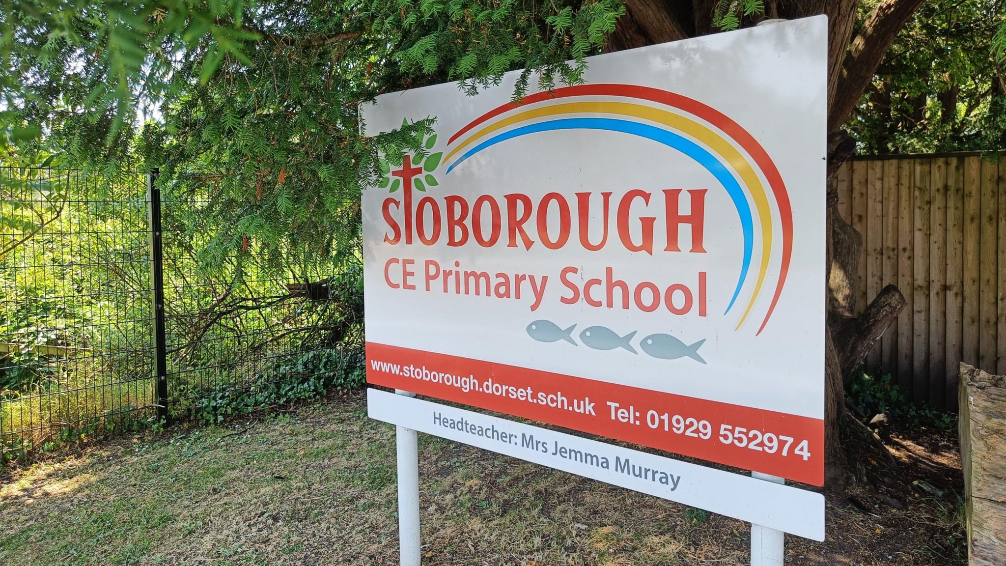 A sign outside the school which is white and red with a rainbow design on. It says Stoborough CE Primary School