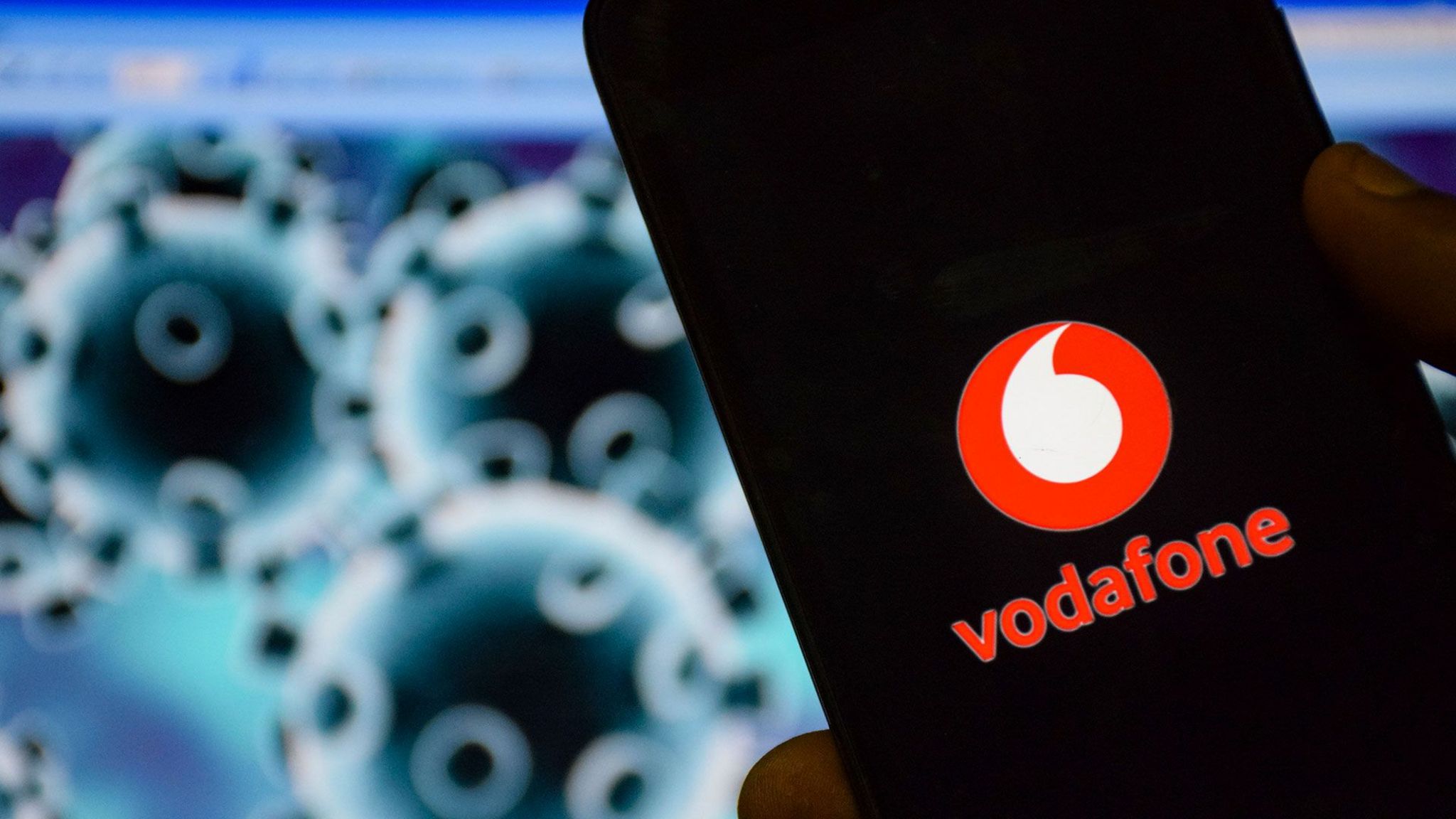 A mobile phone is seen with the Vodafone logo on it in front of an illustration of the coronavirus