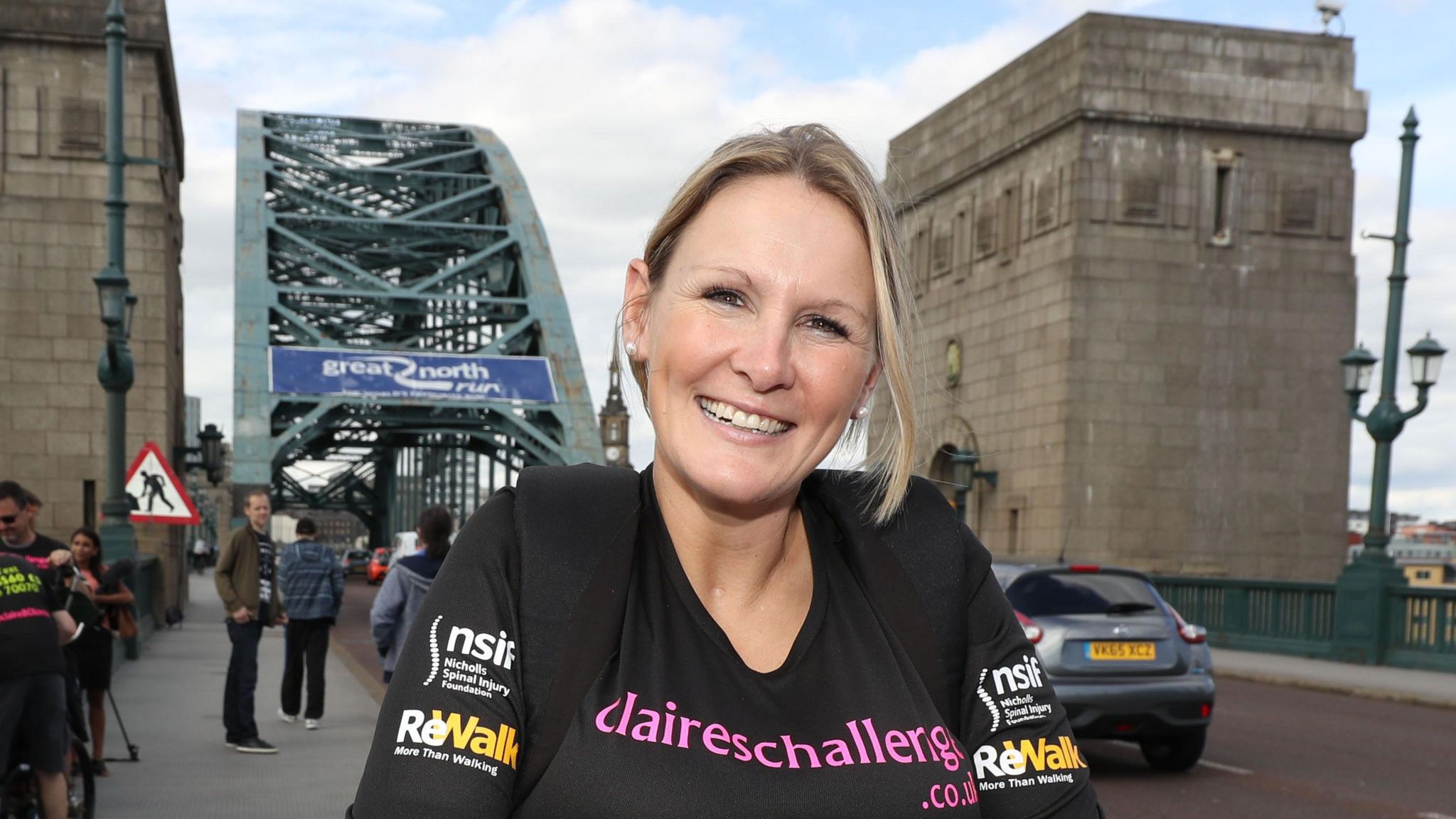 Claire Lomas is pictured after completing the Great North Run. She is wearing a black shirt with sponsor logos. A bridge with the Great North Run logo can be seen in the background.