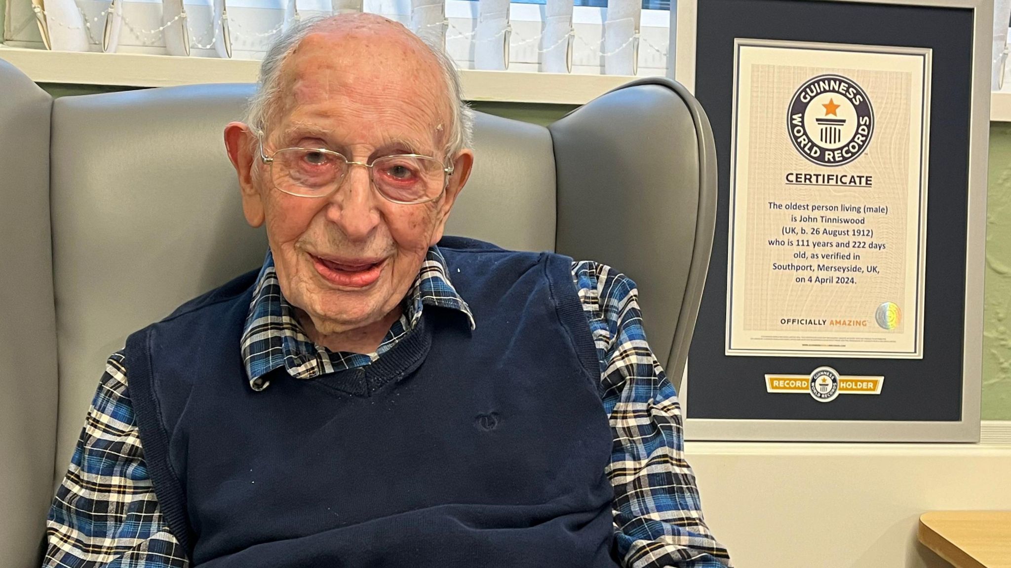 John Tinniswood, with glasses and wearing a blue tank top and blue, black and white checked shirt, sits in a grey chair smiling at the camera. A framed certificate sits on a shelf to his left which shows the Guinness World Records age achievement. 