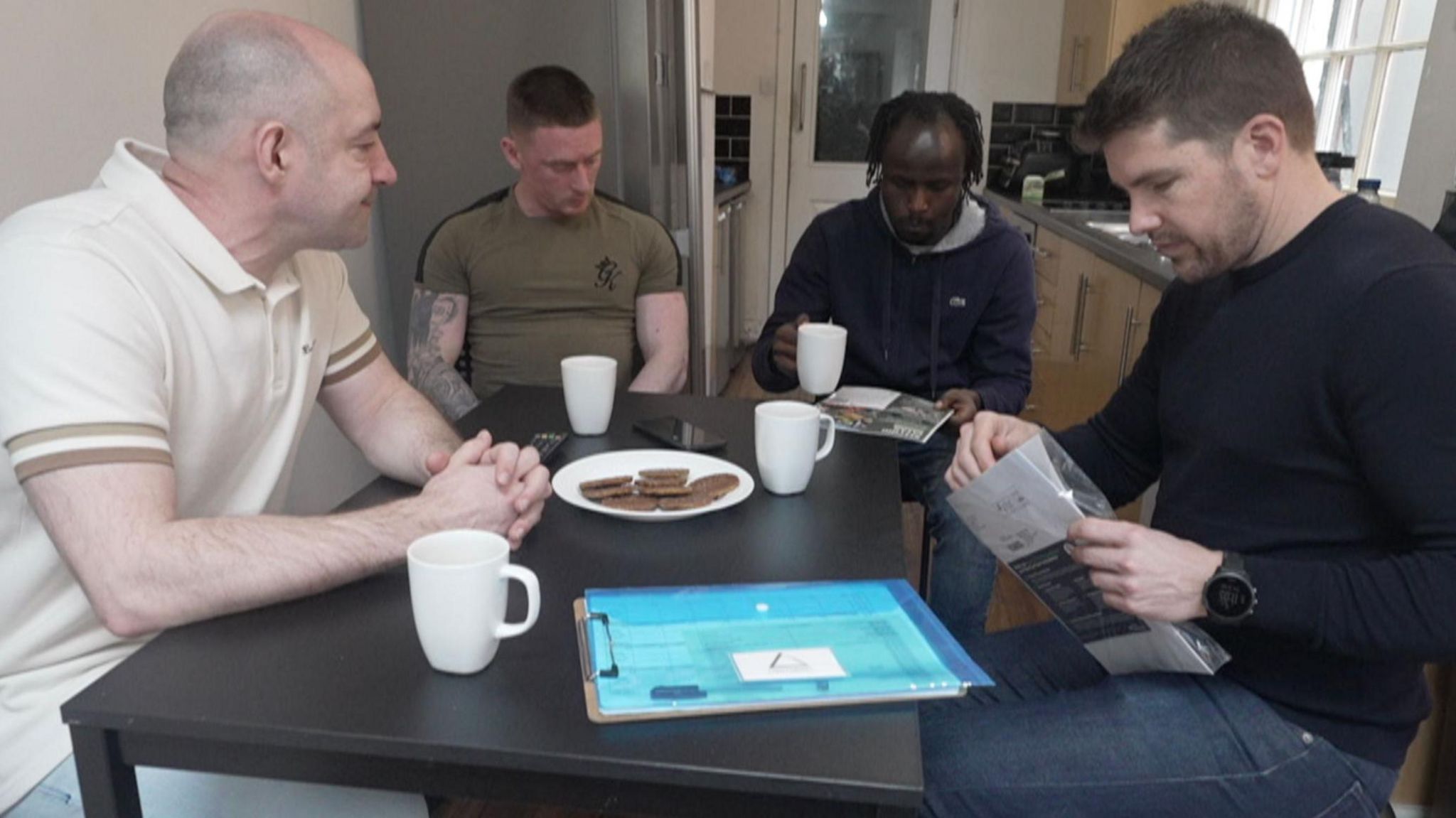 Four men sit around a dining table with cups of tea and biscuits looking at brochures