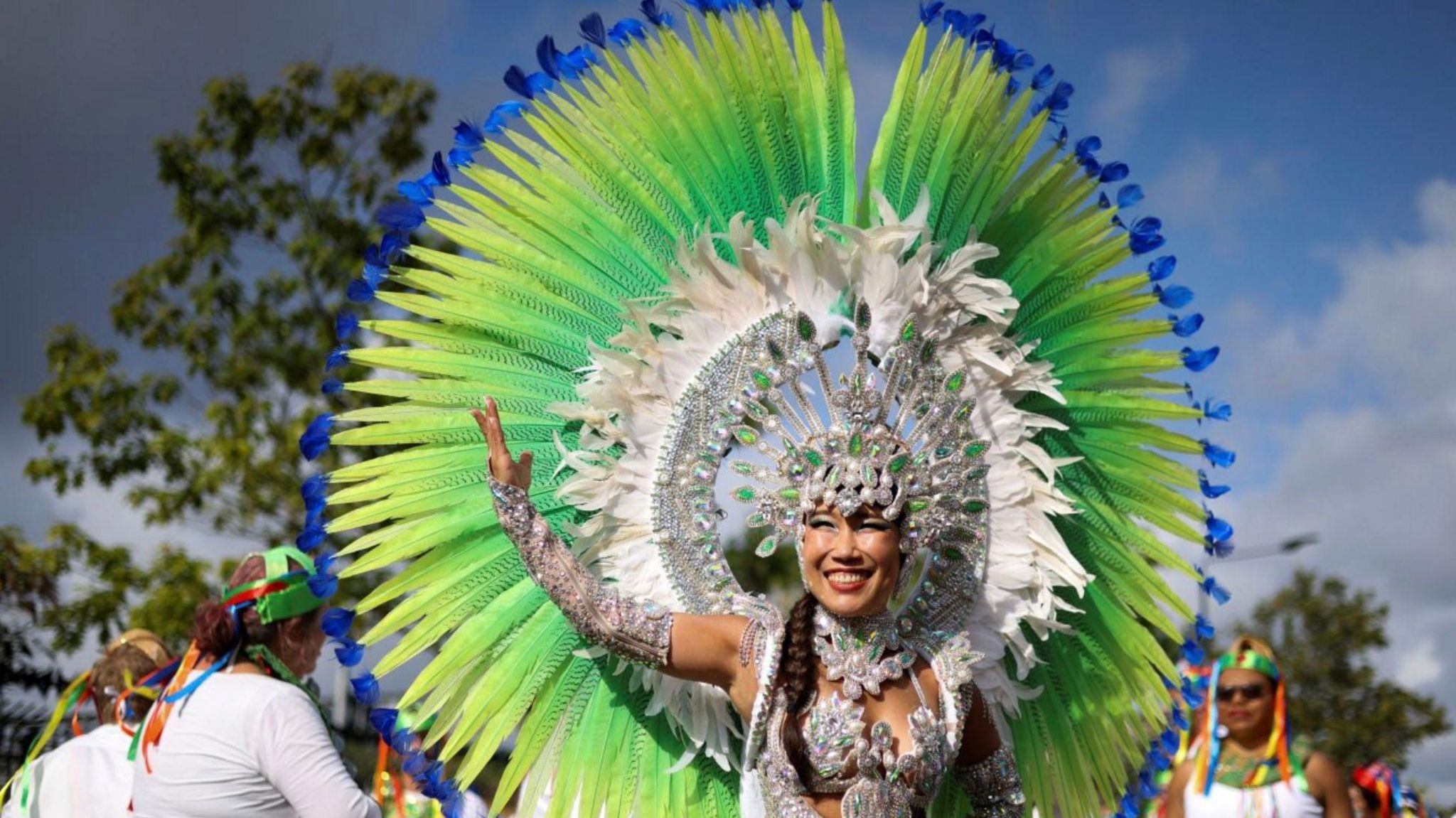 A woman smiles and has her arm in the air. She is wearing an outfit that has large feathers coloured green, yellow and blue attached in a circle behind her with white feathers in another circle within it.