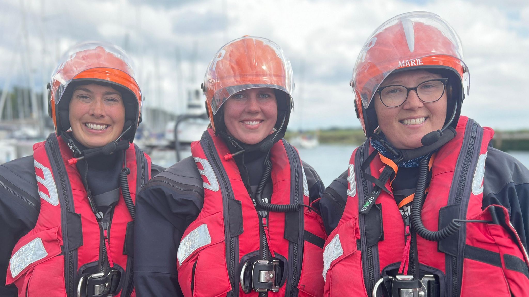 Three smiling female members of the patrol all wearing orange helmets, red life jackets and blue waterproofs. They each are looking directly at the camera, with boats behind them.  