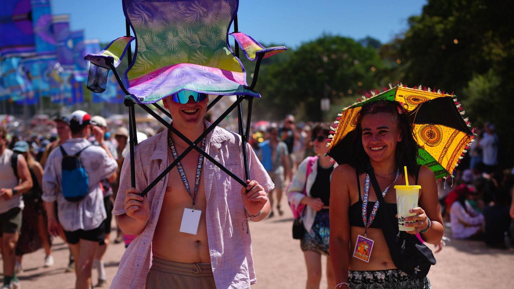 Two people walking through Glastonbury Festival in the sun, a woman has a mini umbrella and a man is holding a camping chair over his head