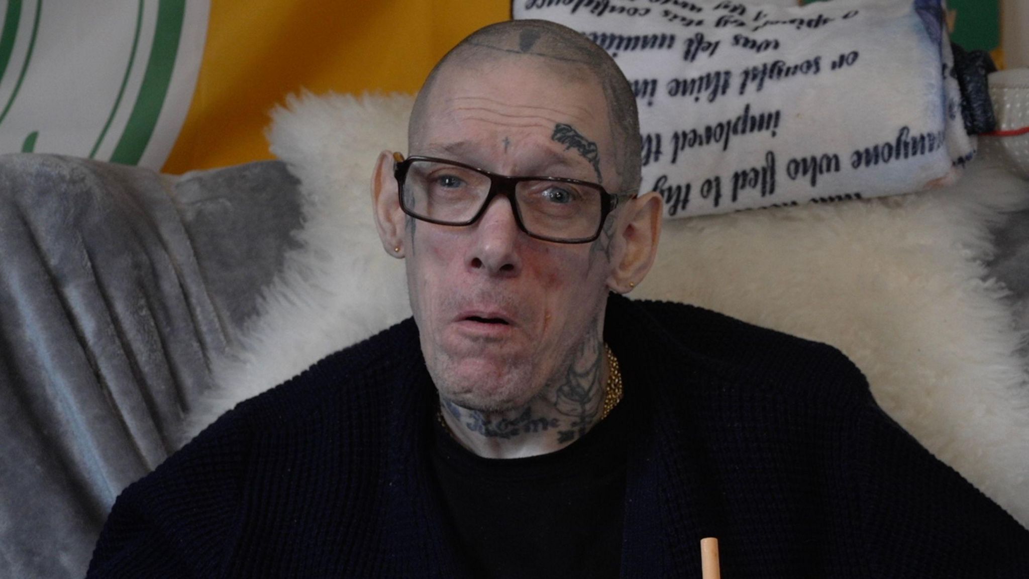 Sam Brackenbury faces the camera wearing a black jumper and black rimmed glasses. He has face and neck tattoos and is sitting in front of a sheepskin throw on a chair. 