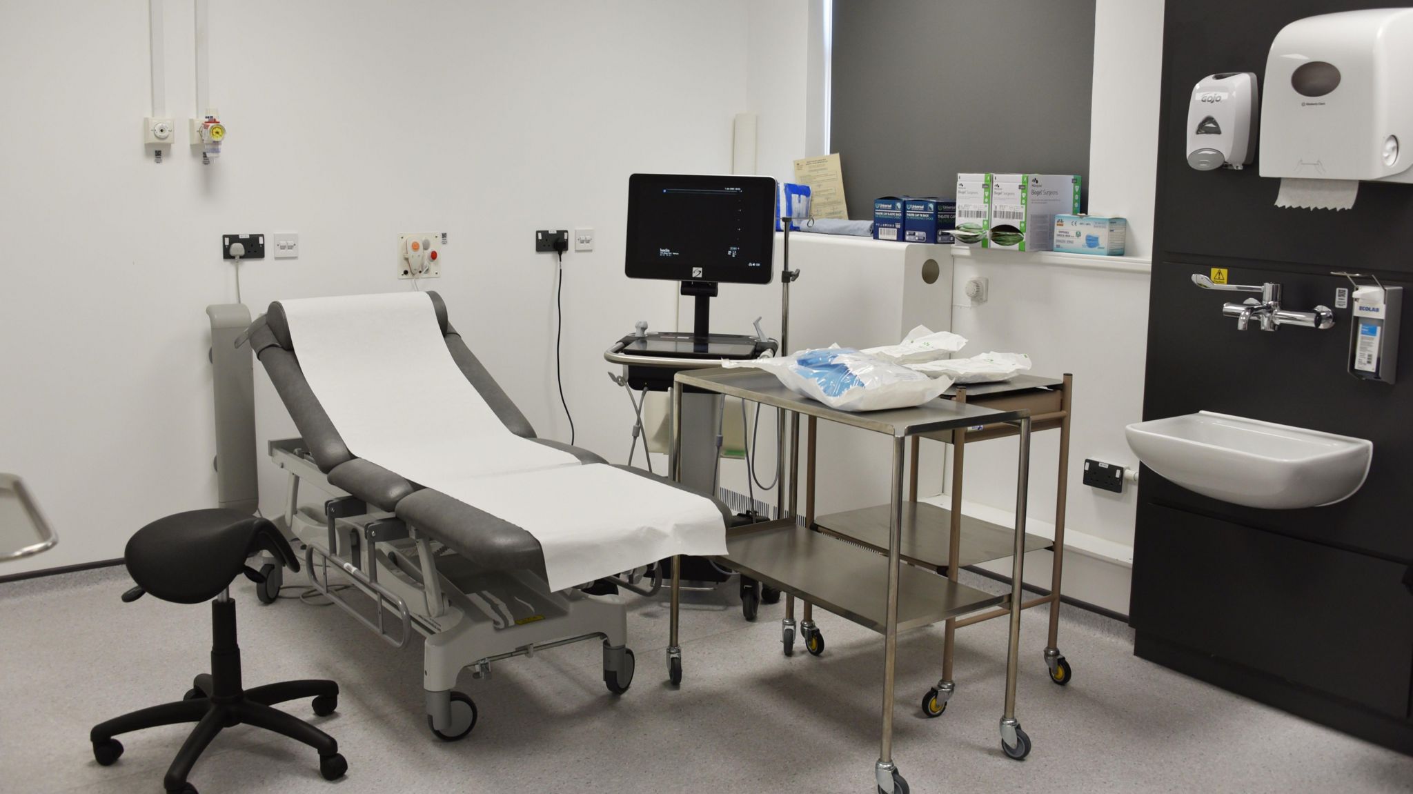 The new treatment room at Musgrove Park Hospital. There is a patient bed covered with disposable paper, beside it a black stool and metal shelves on wheels. There is also a sink to the right of the room, and an ultrasound machine used for the procedure. 