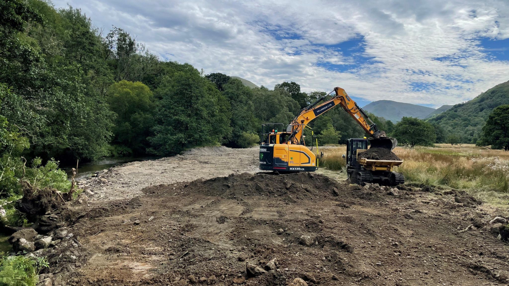 A digger on the banks of the river, removing earth and placing it on the back of a dump truck.