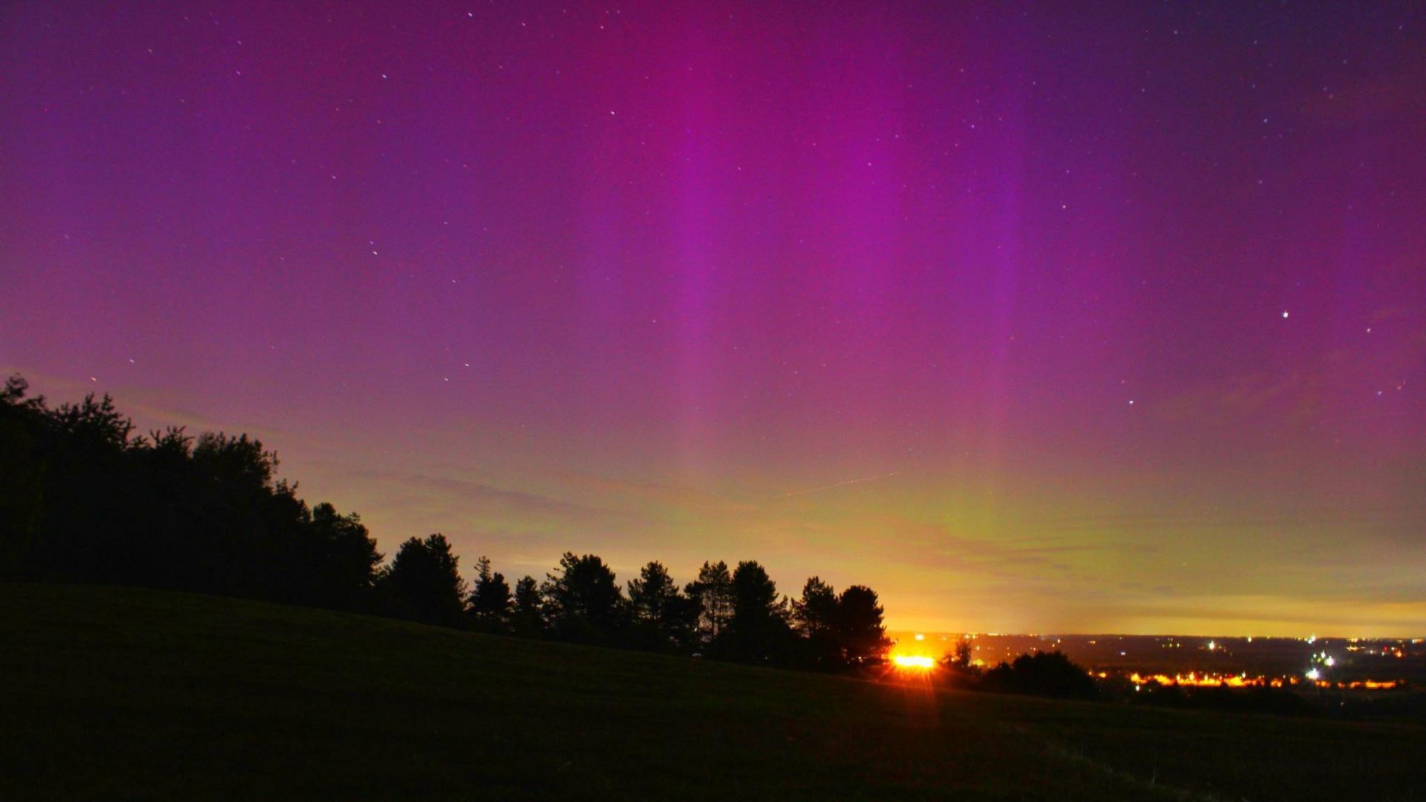 A purple sky with lights from a town visible in the distance and a line of trees silhouetted in the middle distance