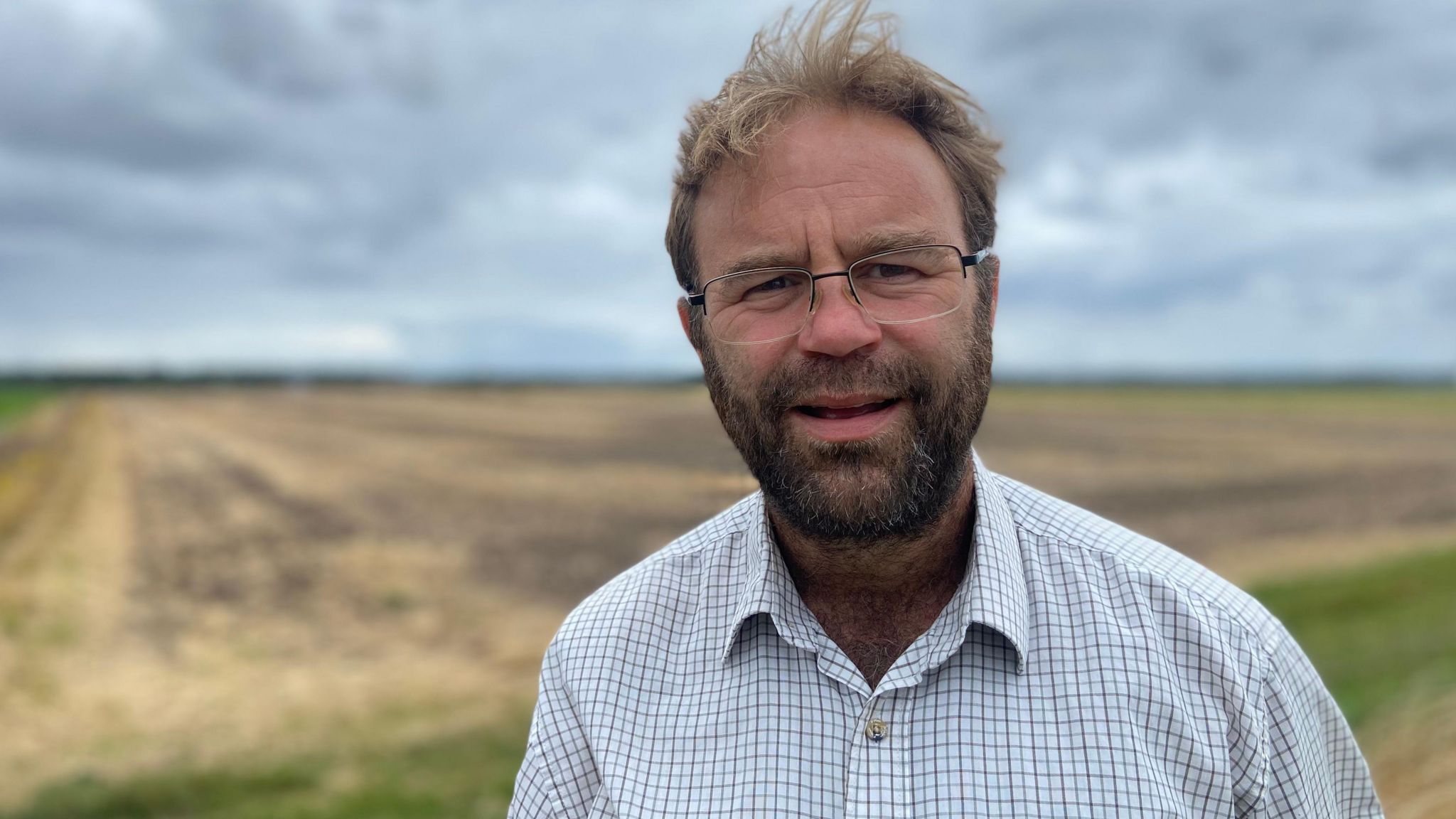 Farmer Colin Chappell, who has brown hair and a dark, full beard, wears a chequered shirt and stands in front of a field