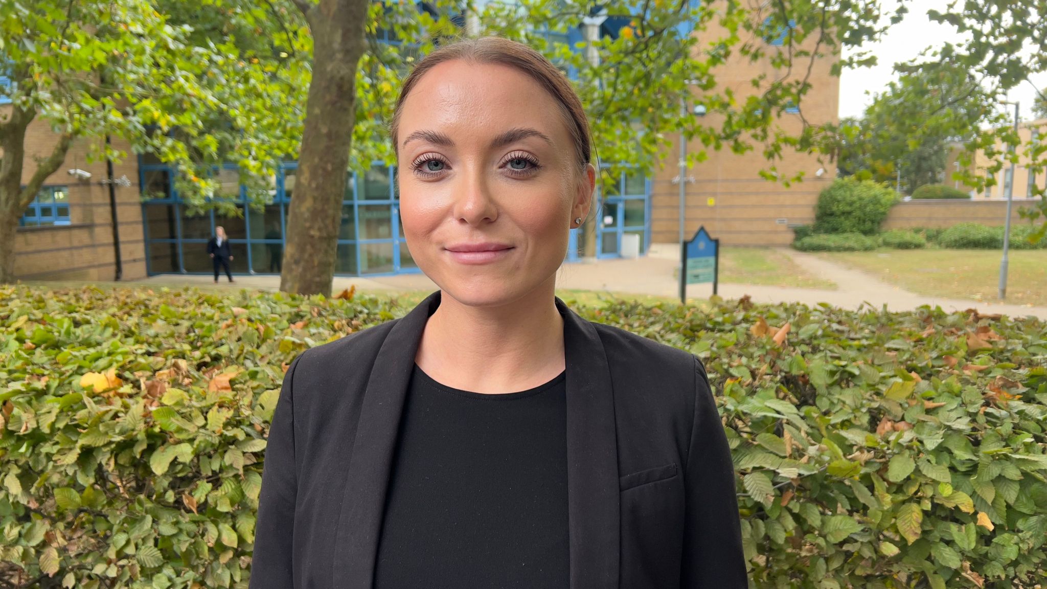 Detective Sergeant Ellie Nudd standing outside Basildon Crown Court. She is looking at the camera while wearing a black top and jacket. Behind her are the courthouse, trees and bushes.