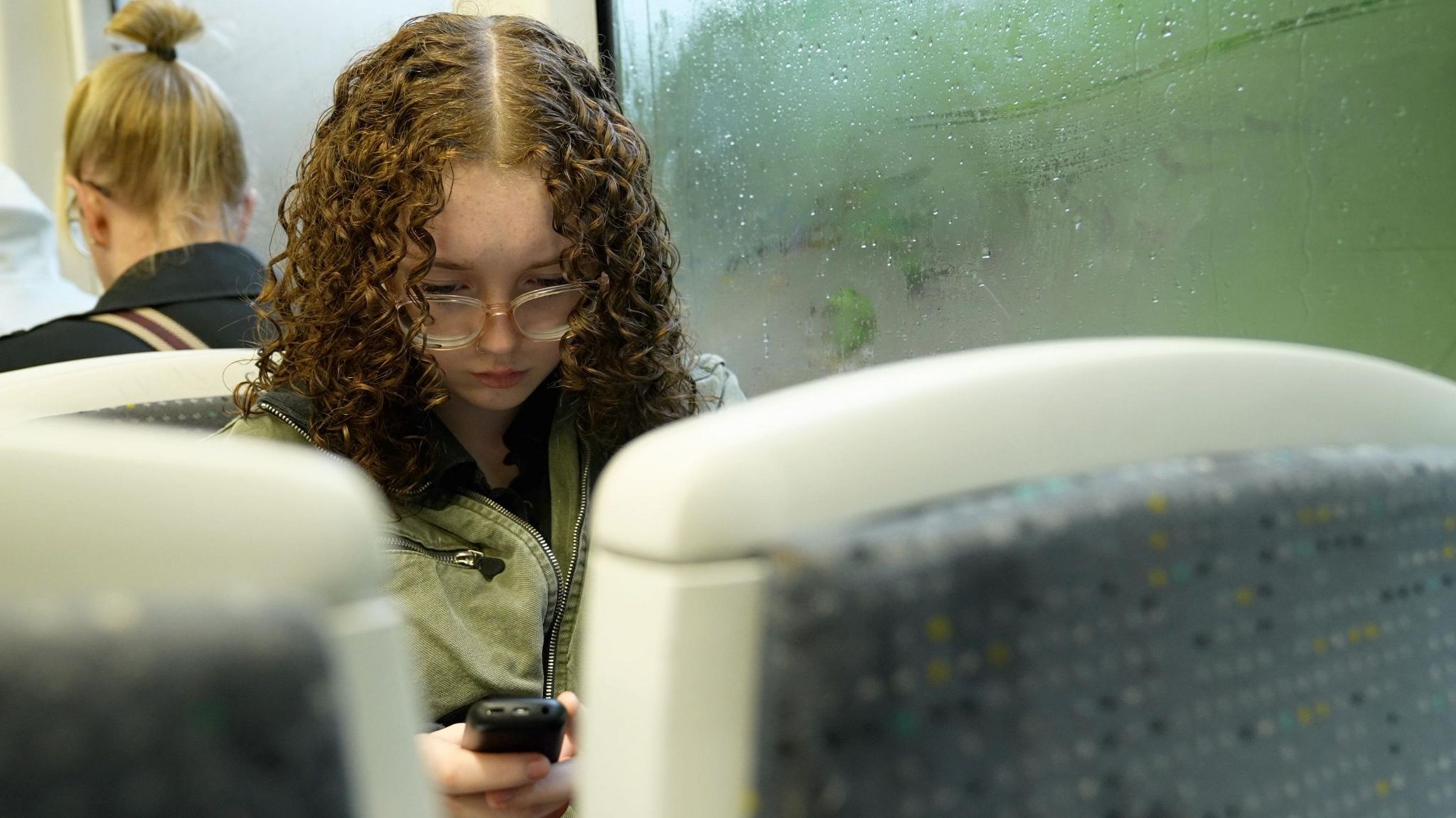 Ruby, wearing a green coat, stares down at her brickphone as she sits on a tram to college