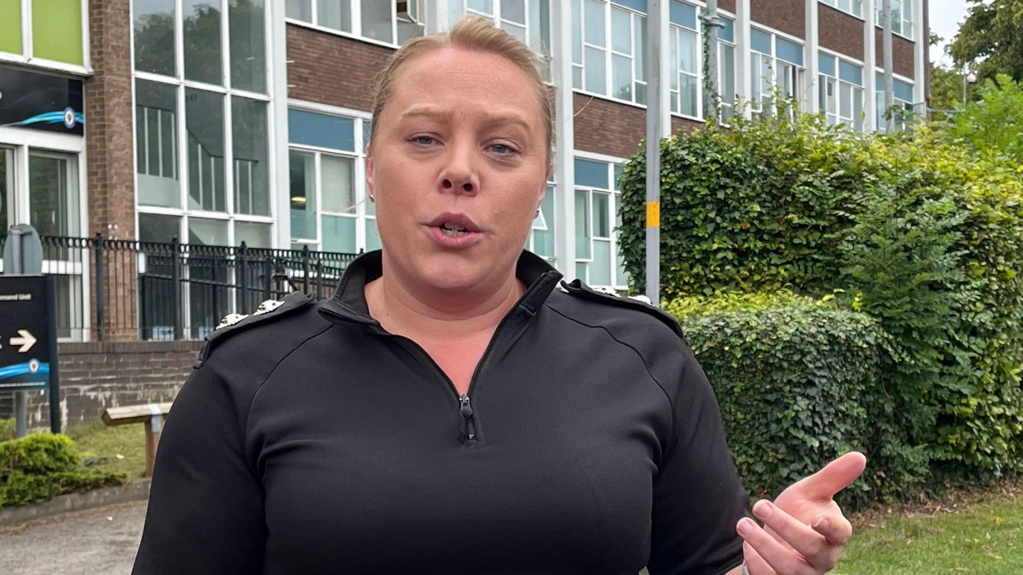 A woman in a black zip up police top. She is standing in front of a police building and some hedges