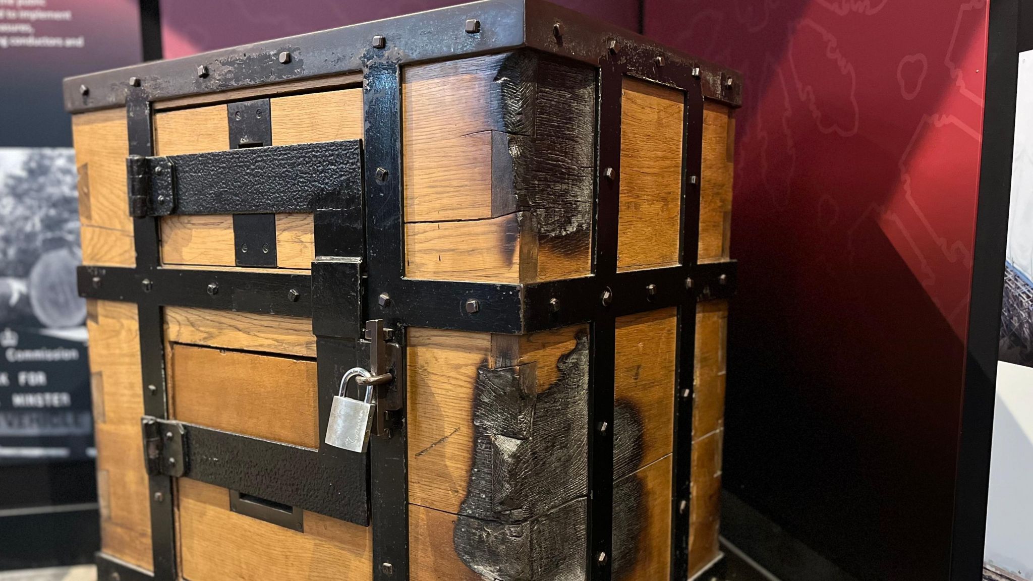 An old donation box with lead bars around it and a big silver lock in the corner of the exhibition showing scorched marks along the front right hand side of the box