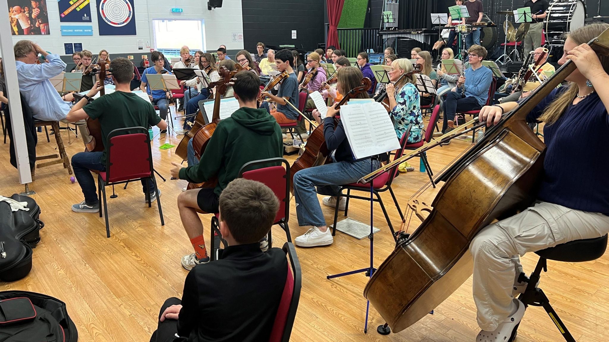 An orchestra sat down in a hall playing together with Elliott in teh foreground with his back to the camera sitting on a chair watching