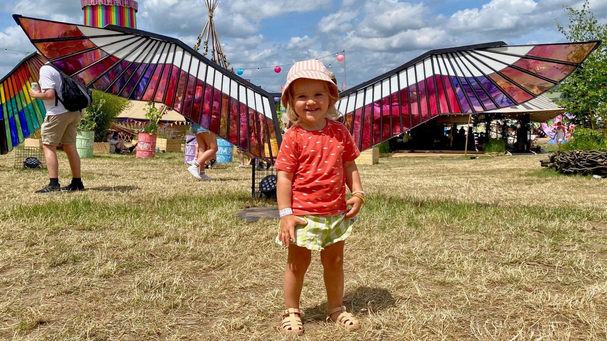 A little girl smiling and standing in front of the pink and purple stained glass wings at Glastonbury 