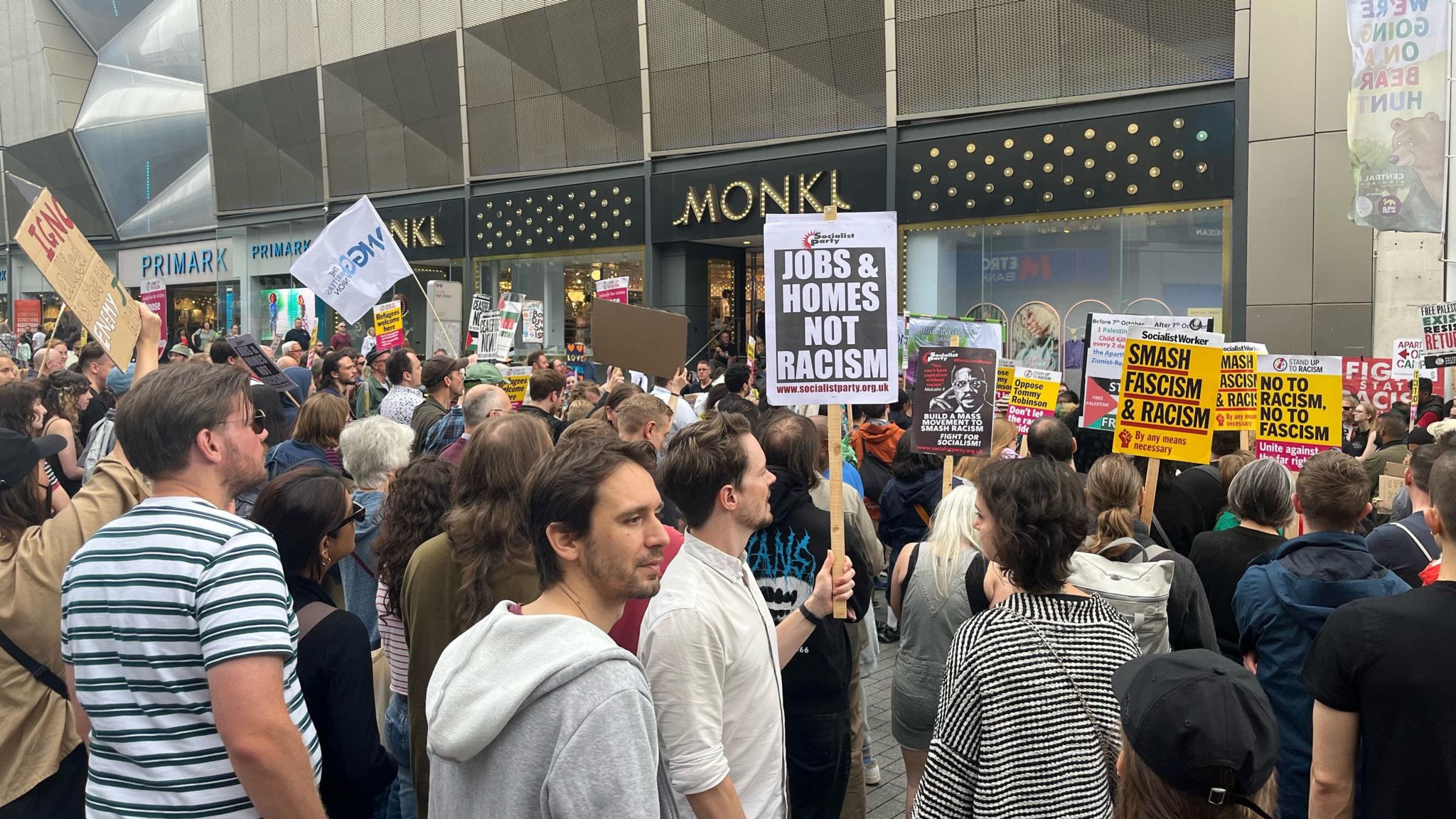 Crowds of people stand outside a row of shops, with several people holding up protest signs such as 'Jobs & Homes Not Racism'