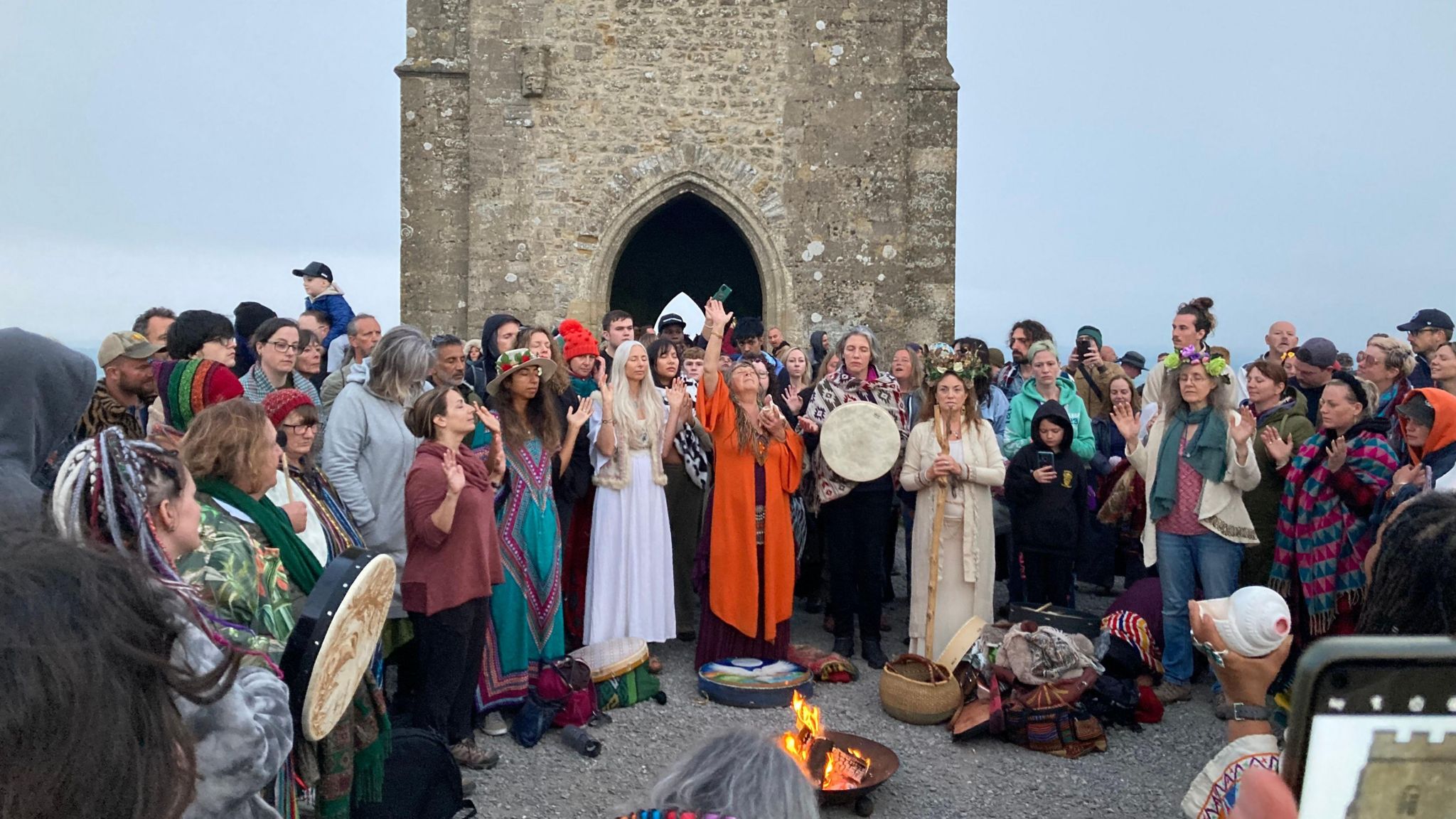 A crowd gathering at the Glastonbury Tor around a fire as dawn breaks