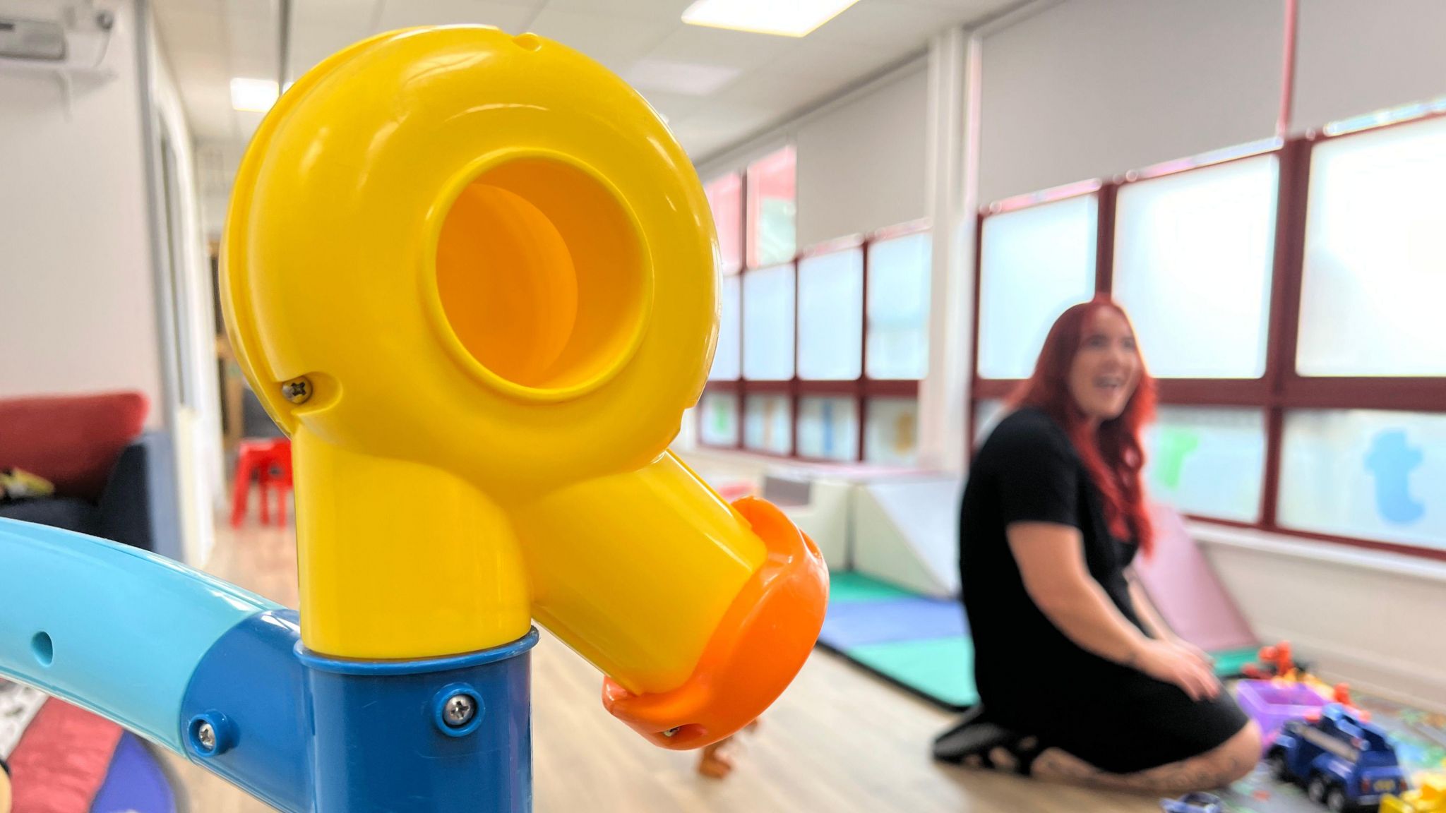 A large plastic yellow toy in an abstract shape is visible in focus in the foreground. A blurred woman with long red hair, wearing black, can be seen in the background kneeling with toys in front of her. 