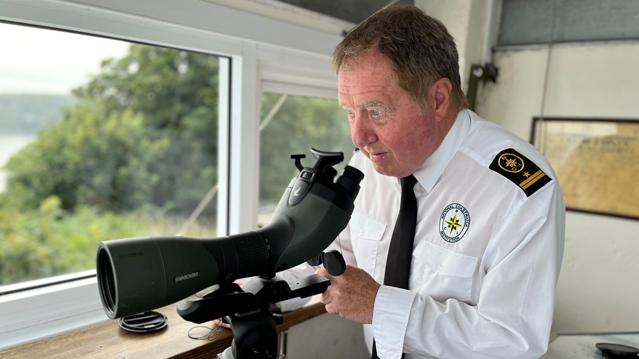 Andy Westward, station manager of National Coastwatch station at Froward Point, dressed in a white shirt with epaulettes and wearing a black tie, looks out of a window with a telescope