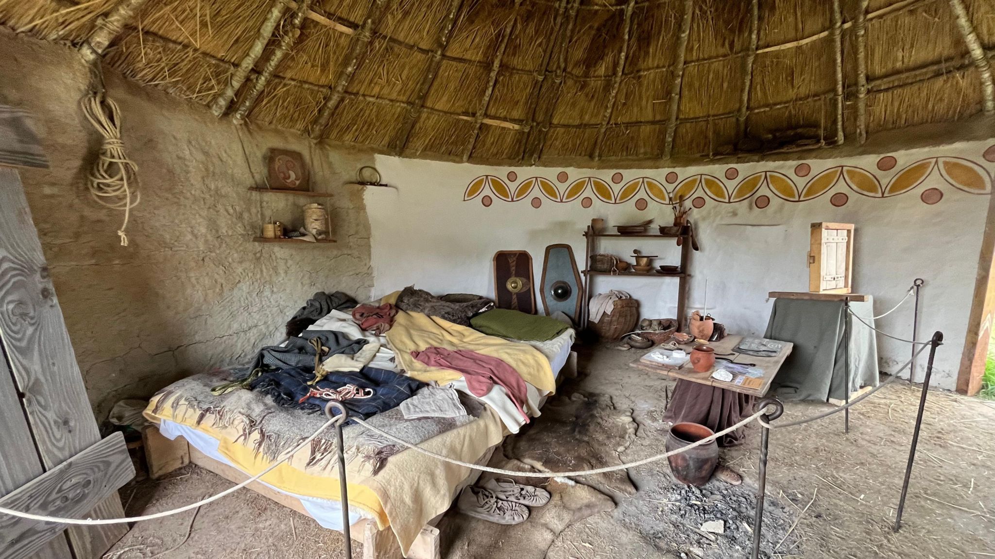 The inside of a small round building with a thatched roof, half mud walls and have painted white with a little decoration. Objects include a bed, animals skins and pots.