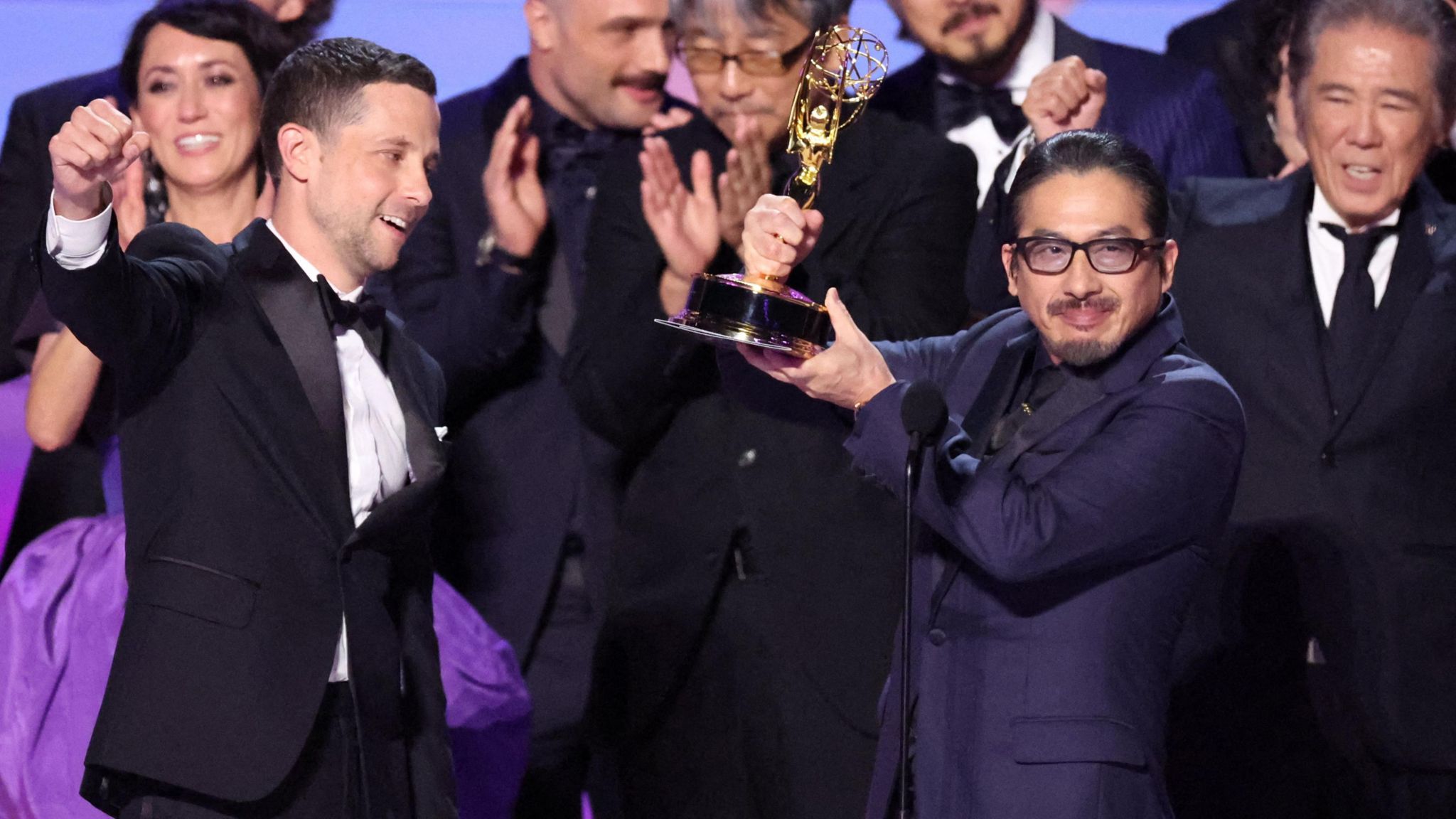 Shogun's Hiroyuki Sanada holding an Emmy aloft on stage, surrounded by with the show's other stars and makers 