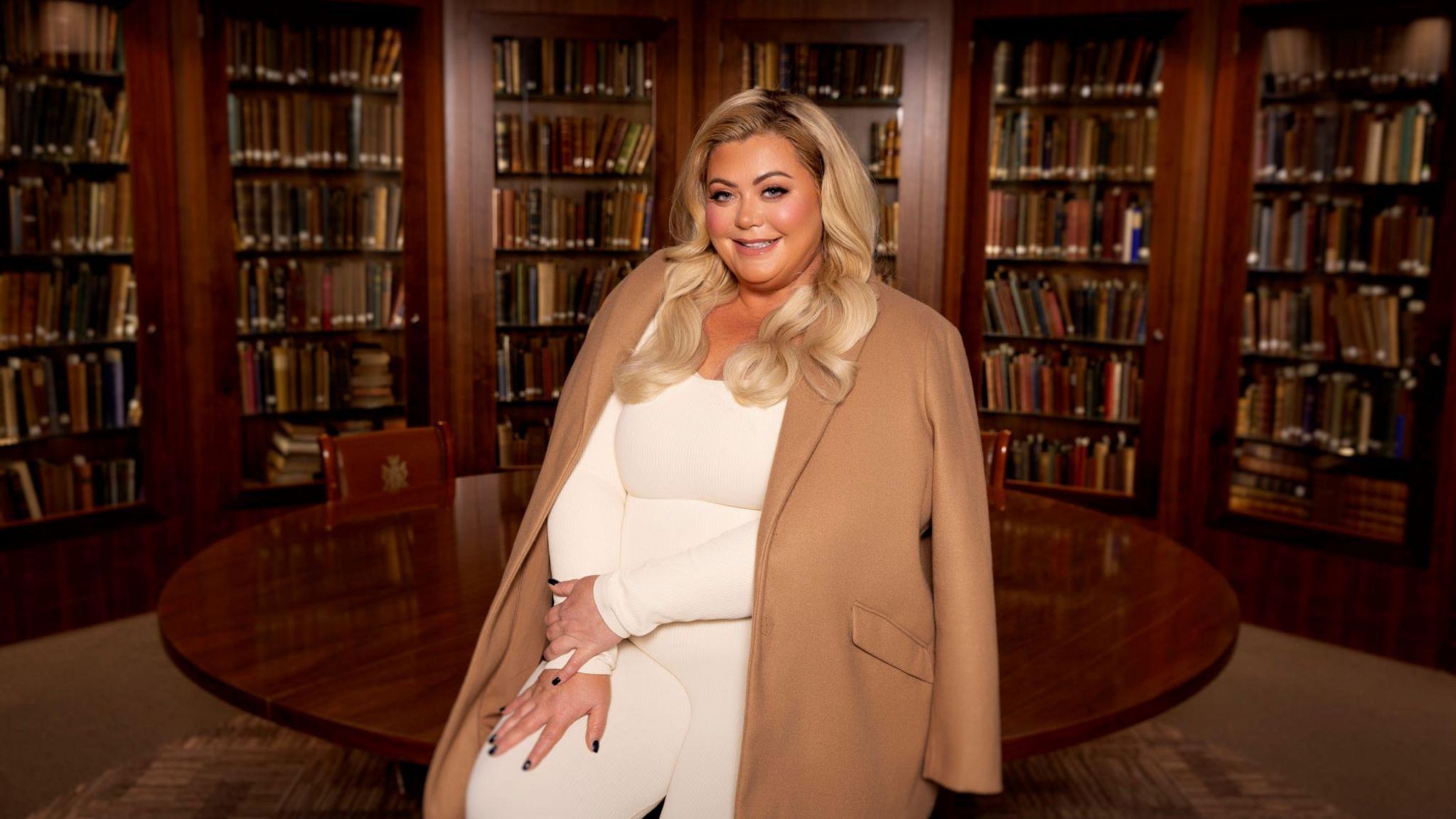 Gemma Collins is wearing a white bodysuit with a cream coat draped over her shoulders. She is smiling at the camera with her hands on her leg. The room she is in has a large round table and six tall bookshelves.