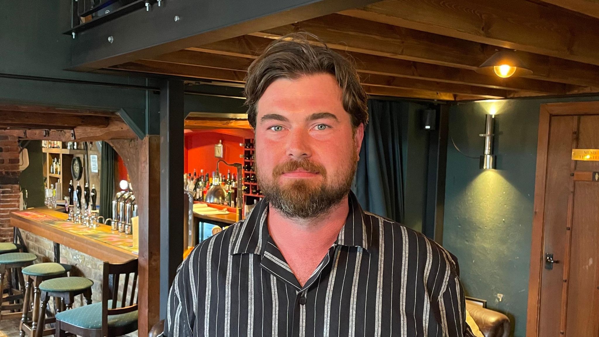 Rory MacRae, looking directly to camera, wearing a striped black and grey shirt and short-trimmed beard, stands near the bar in The Bull pub. The walls are dark green and a wooden beam can be seen over the bar area, which has hand-pumps for ales and bar stools next to it.