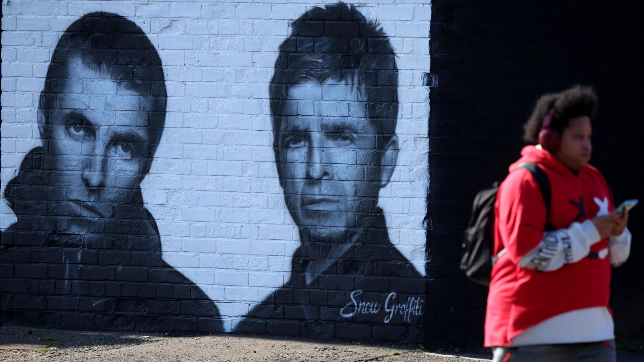 A man with headphone and a red jumper walks past a black and white mural of Oasis on a brick wall. 