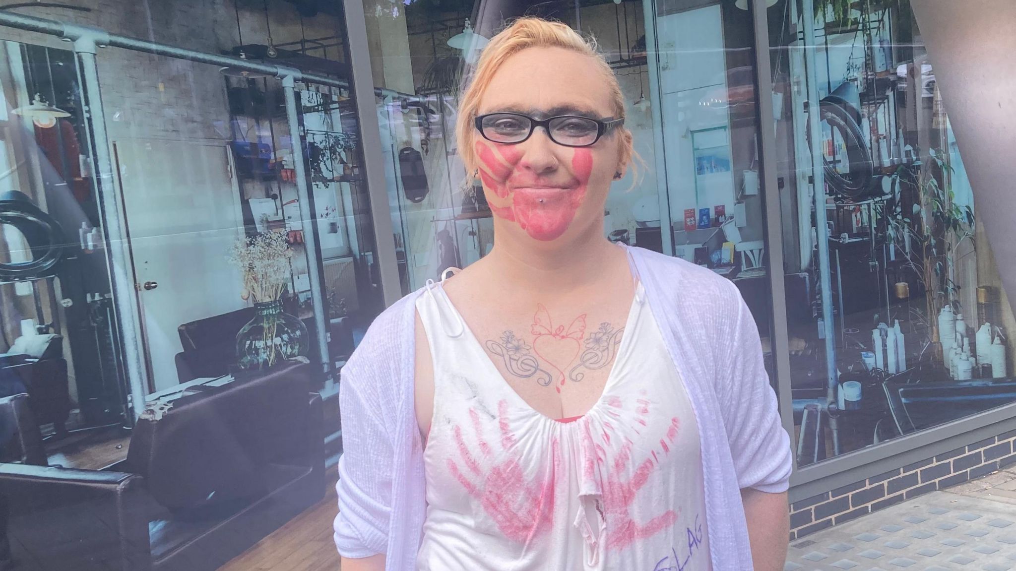 A blonde woman wearing glasses with a line of black face paint covering her eyes and a red hand mark over her mouth. She also has two red handprints on her top over her breasts.