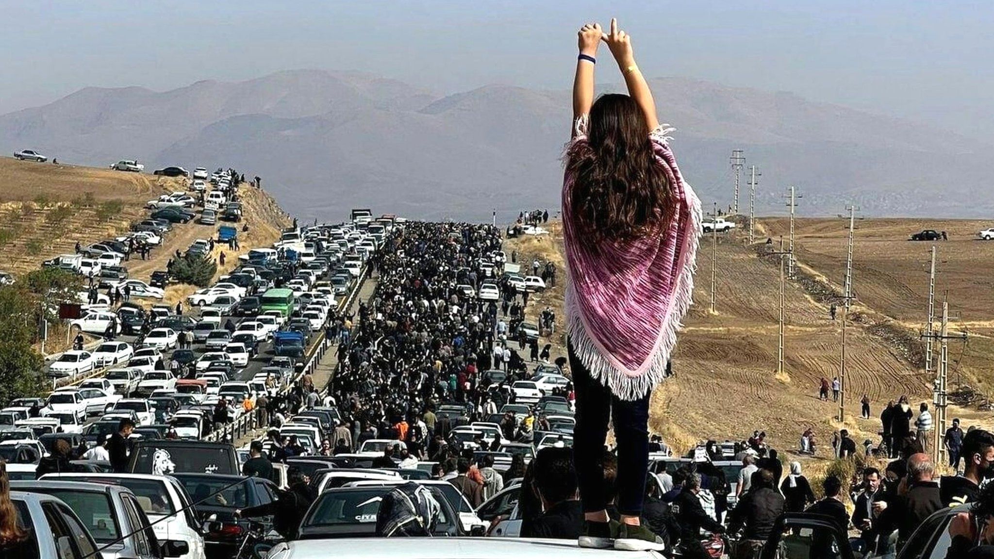 A young woman without a hijab stands on a car as a huge crowd walks towards the Aichi cemetery in Saqqez, Iran, to visit Mahsa Amini's grave on 26 October 2022
