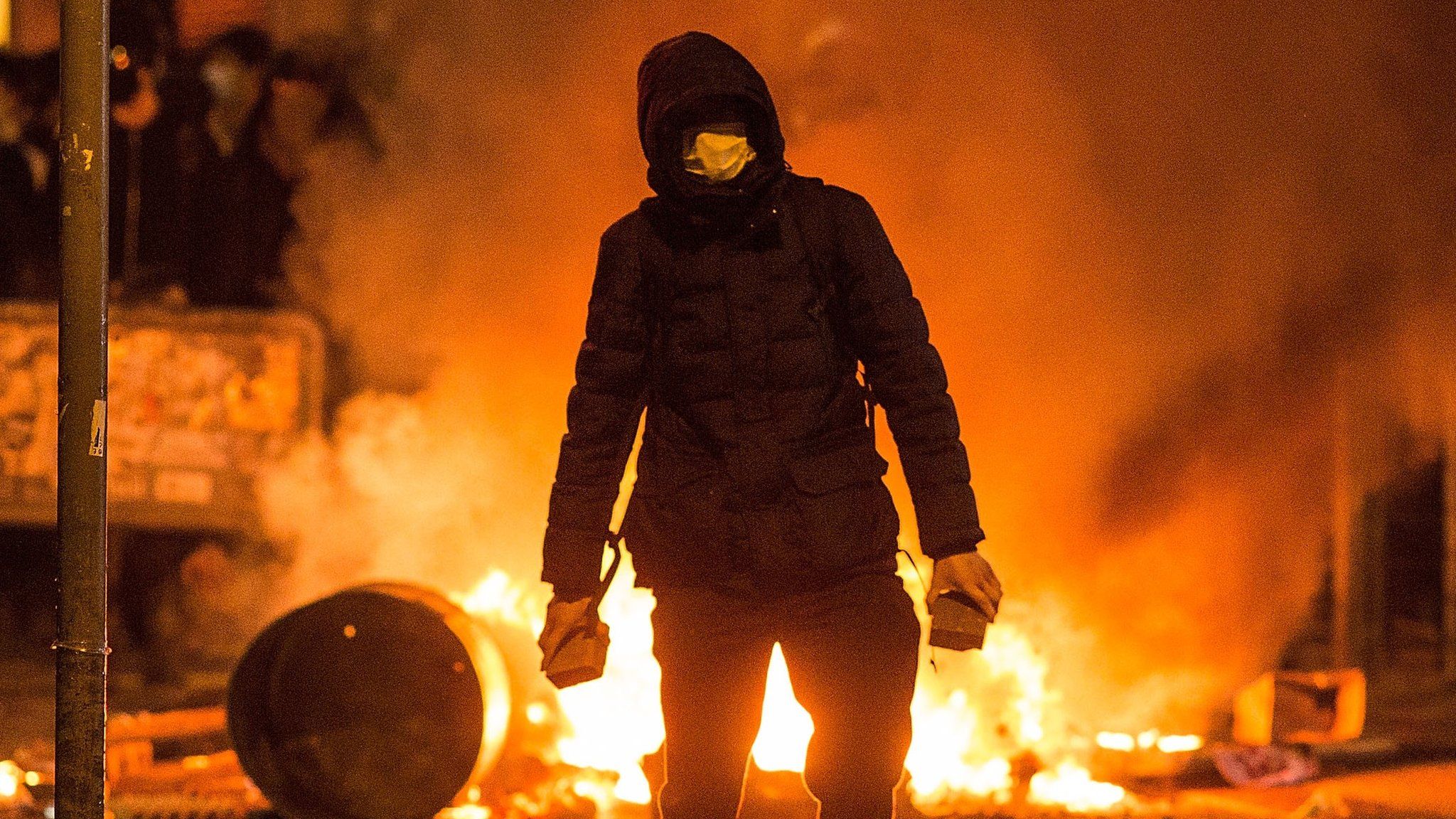 A rioter carries bricks, in front of a burning barricade