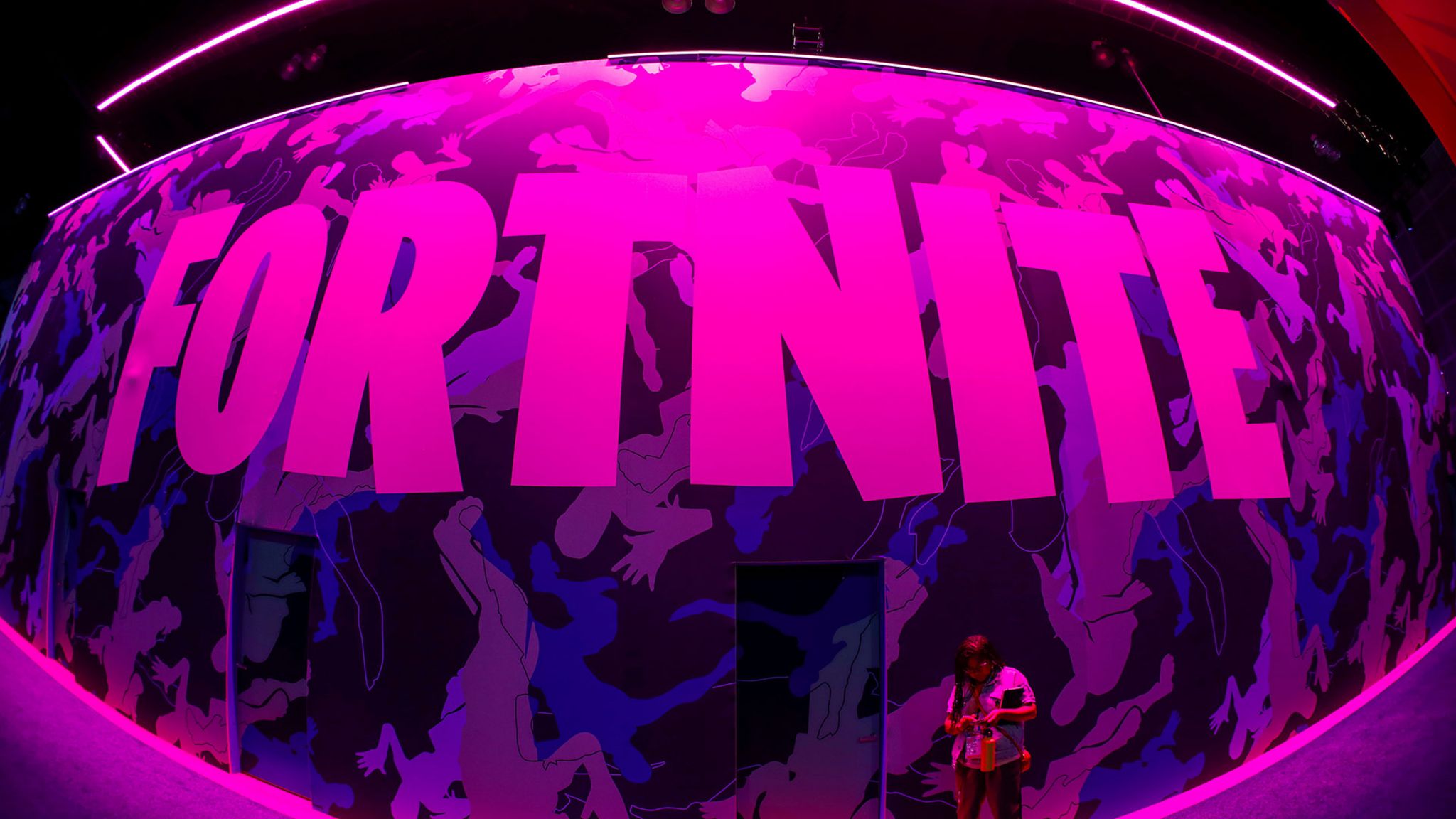 A giant Fortnite logo is seen here at an event, lit by purple lights with a bystander in front of it, appearing small by comparison to the sign's oversized scale