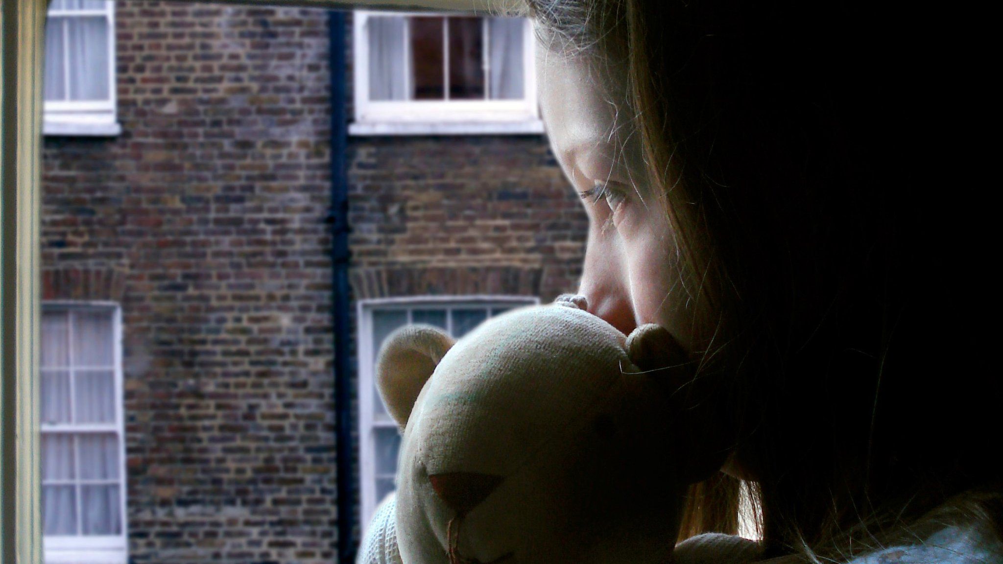 Young girl looking out of a window holding a teddy bear.