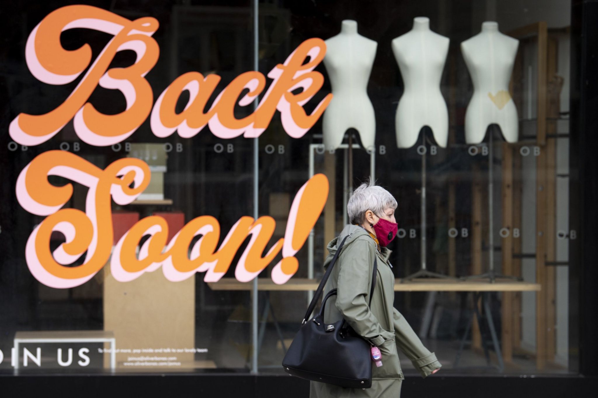 Woman in mask walks past closed shop with the words 'Back Soon!' on the window