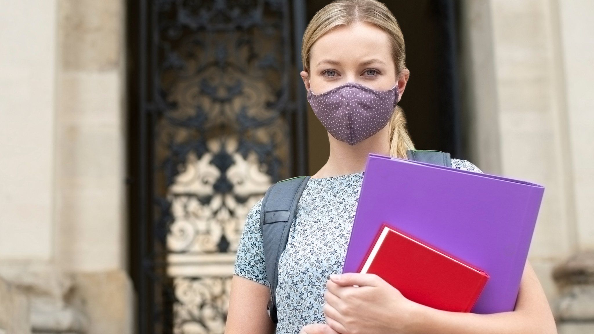 A woman wears a mask while holding folders outside a university building