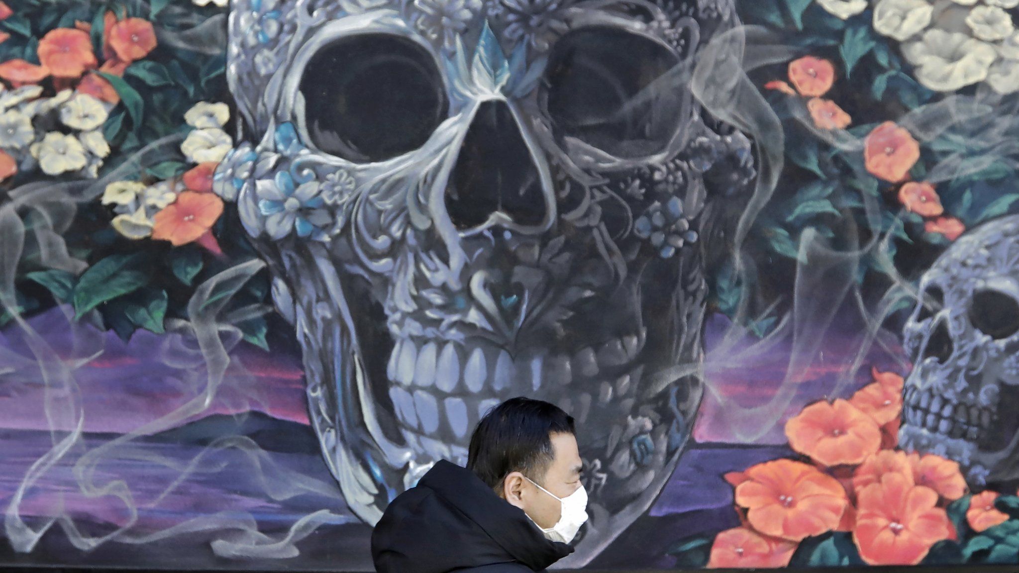 A man wears a protective face mask as he walks in front of a mural of skulls and flowers in New York