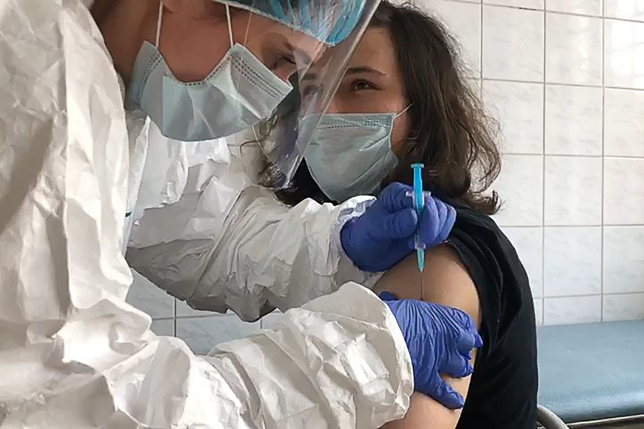 Женщине в маске врач вводит вакцину в полных СИЗ и с козырьком