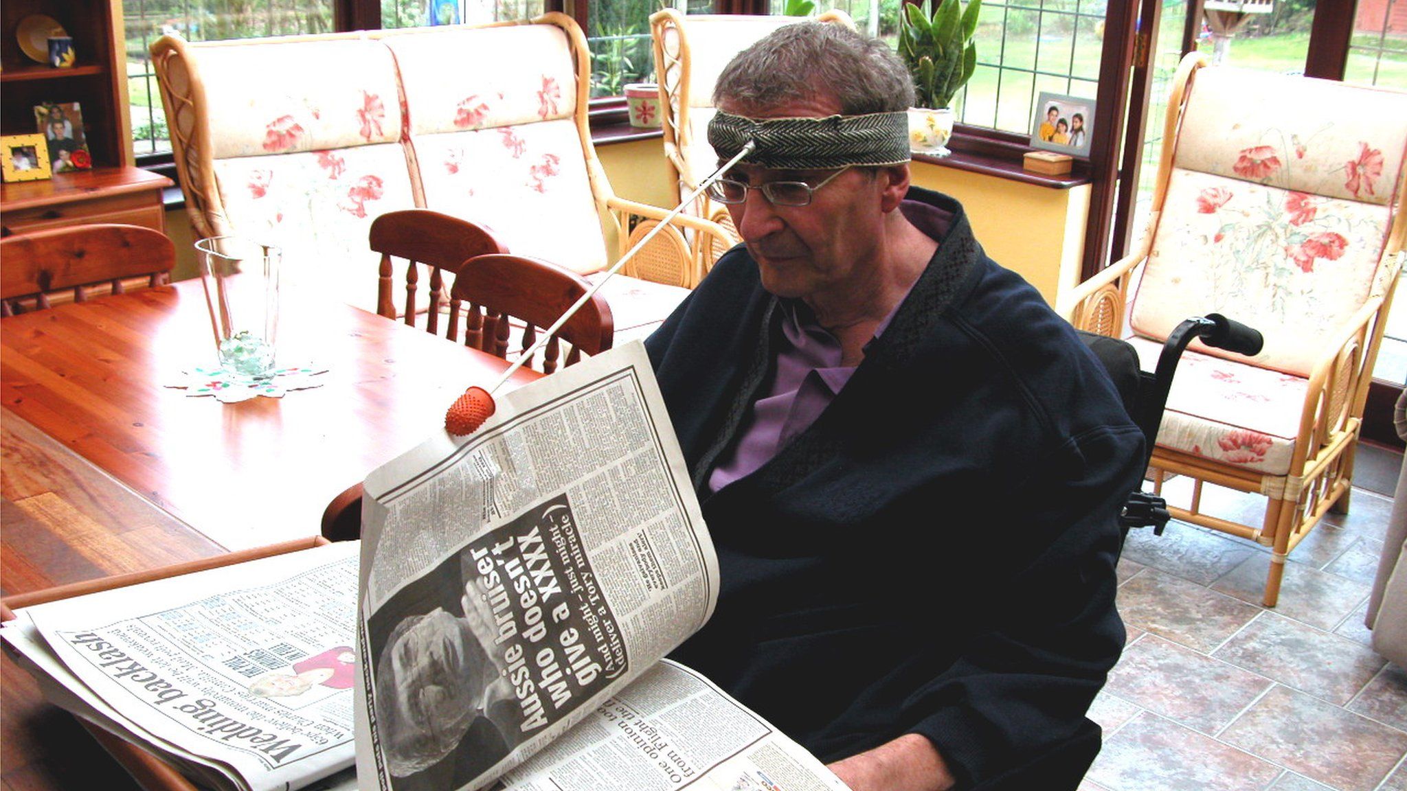 A man turns the page of a newspaper using a modified head-stick with a rubber attachment to grab the paper.