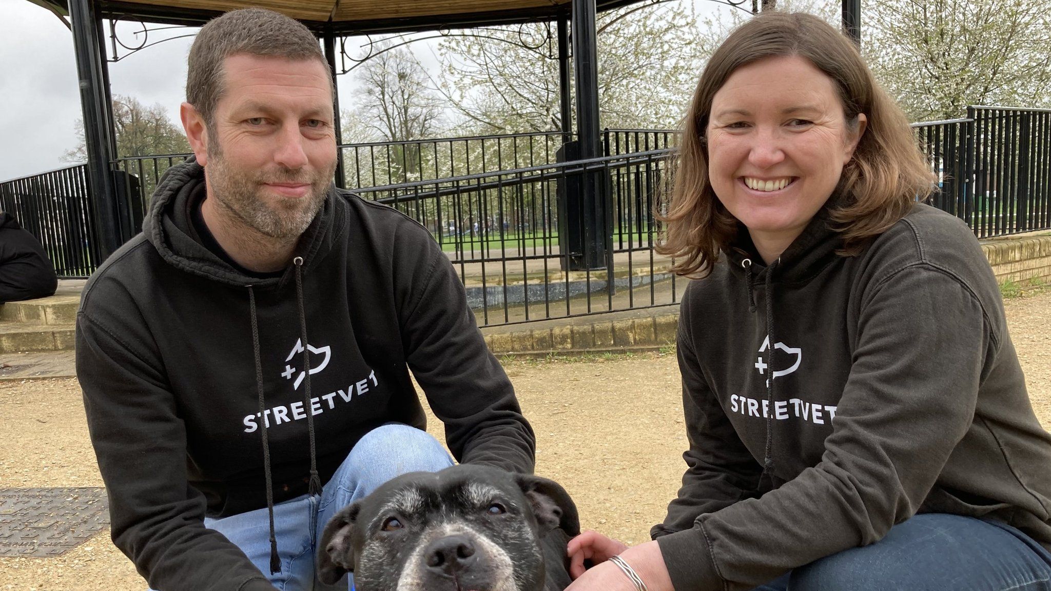 Two volunteer vets knelt down with a black Staffordshire bull terrier with its tongue poking out