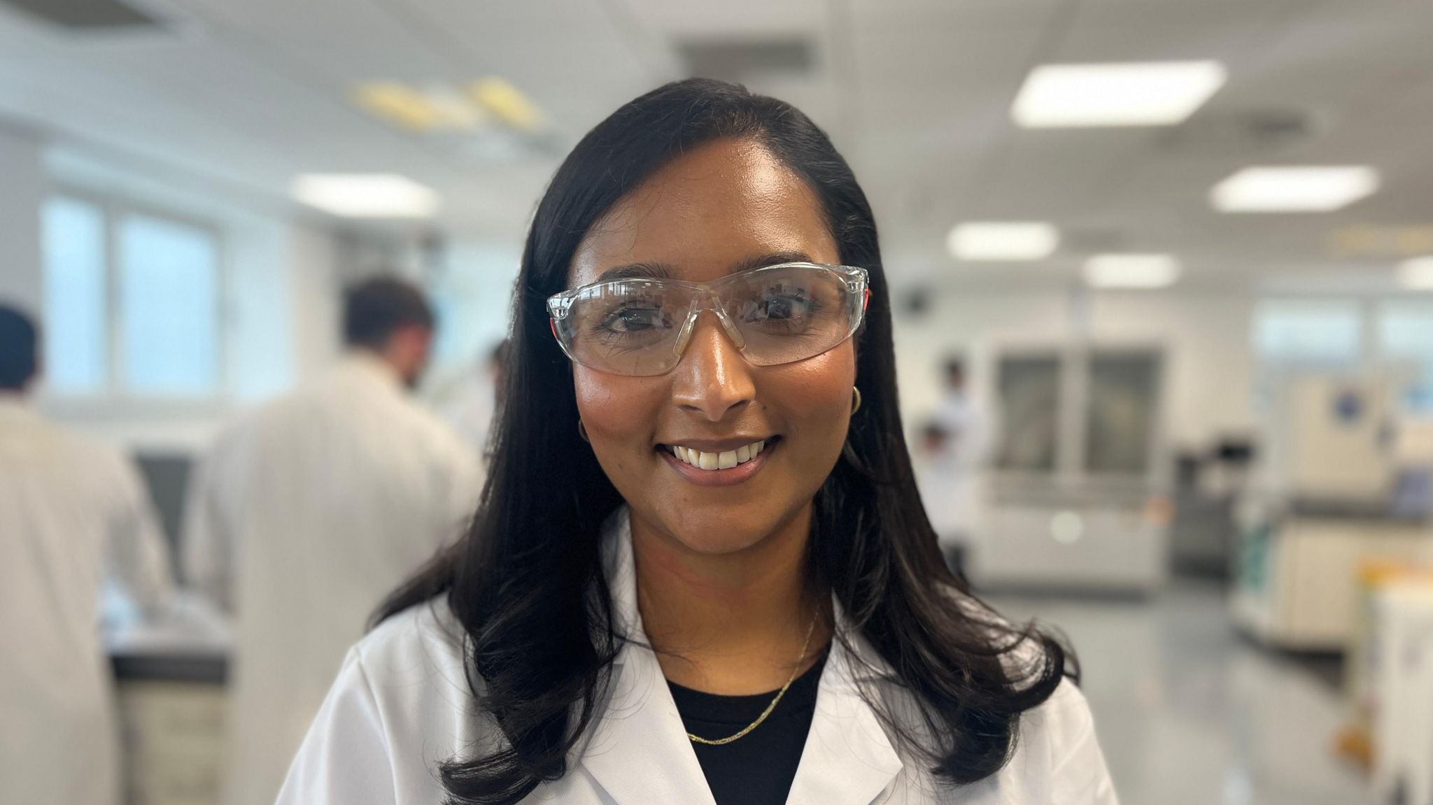 Sabrina tbc wearing a white lab jacket and safety glasses looking toward the camera