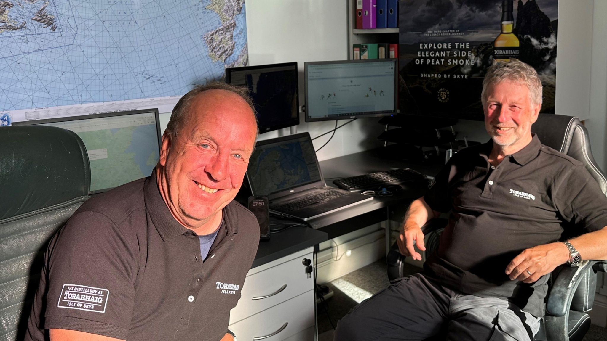 Two men sat in the flight control office next to computers and maps