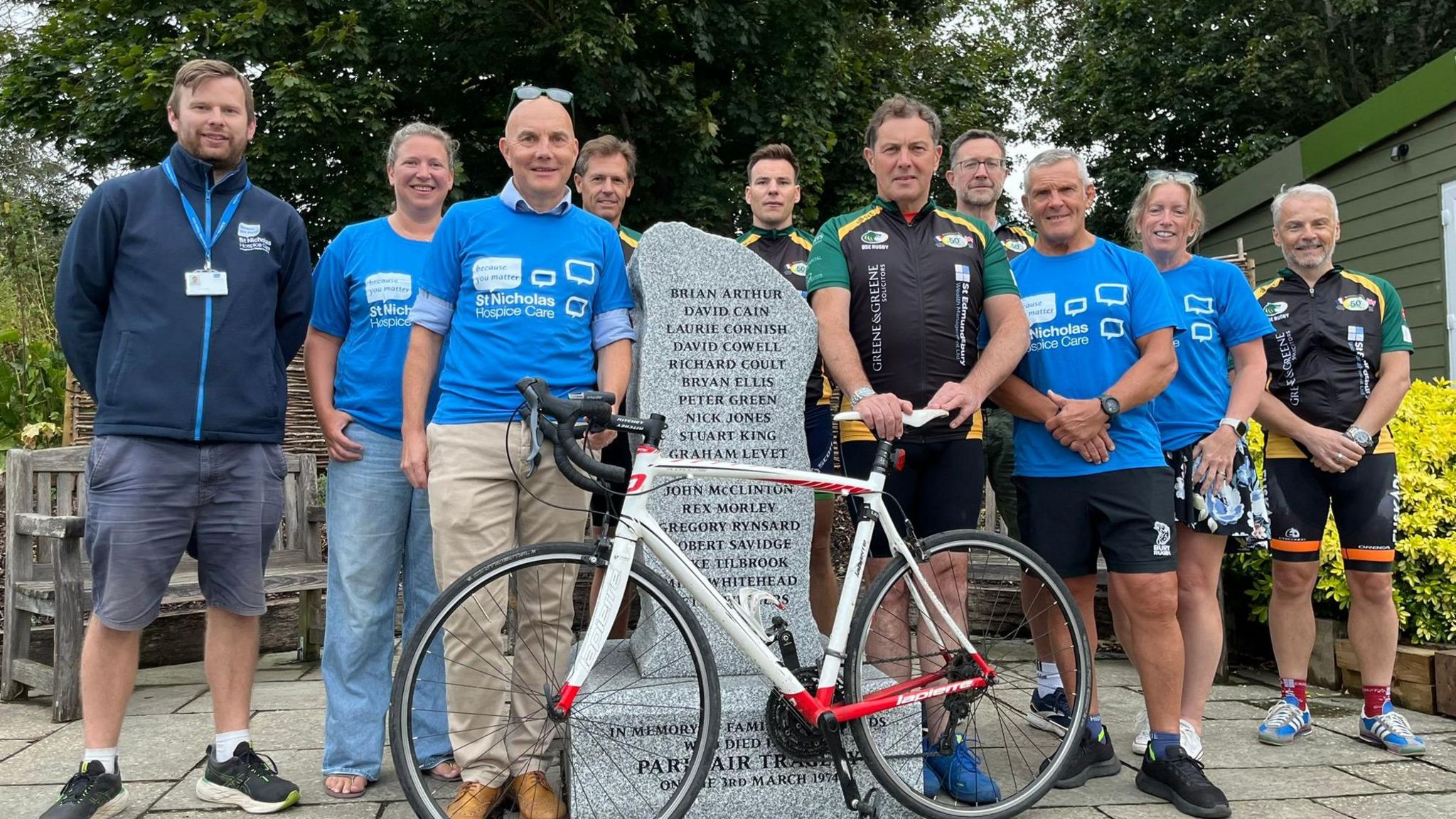 Cyclists stand in front of a stone memorial at Bury Rugby Club. Many are wearing blue t-shirts for St Nicholas Hospice, a charity benefitting from the ride. Others are in black and green cycling shorts and tshirt. The stone has the names of victims etched into it and a bike is being held in front. 