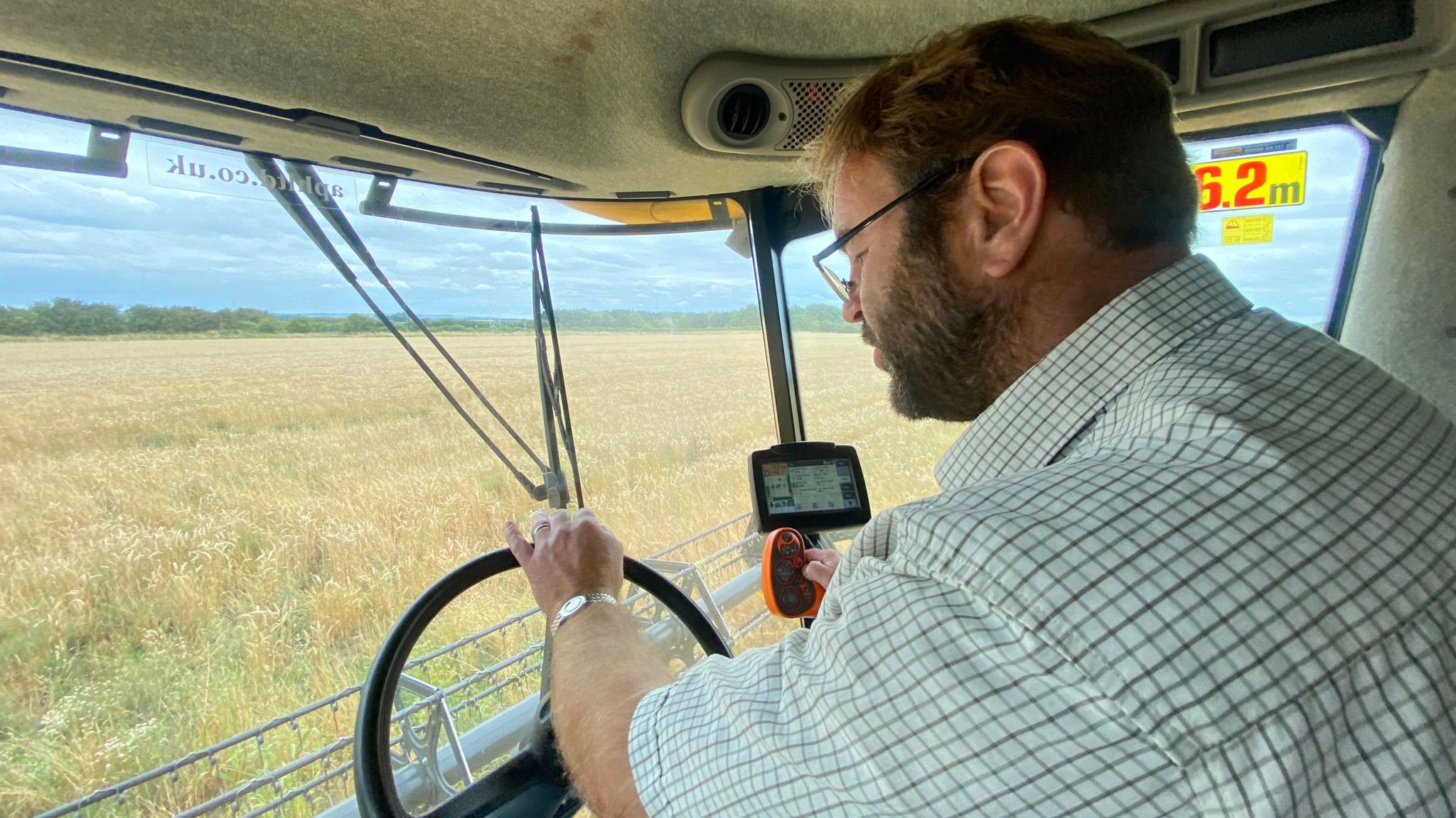 Colin in a chequered shirt in the cab of a combine harvester, harvesting a patchy field of wheat