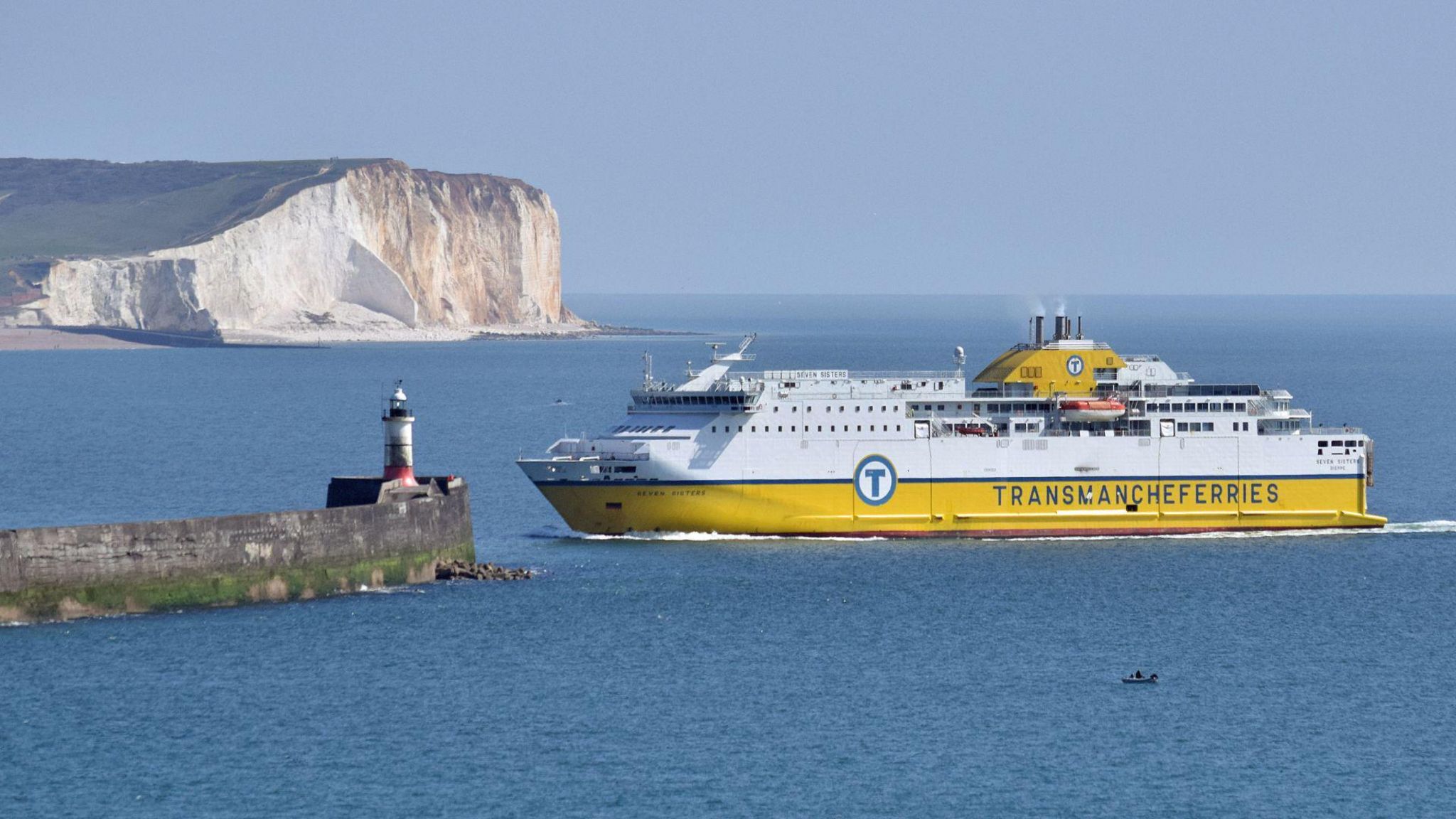 A modern DFDS ferry arriving into Newhaven Port. The top half of the ferry is white, the bottom is yellow. It is travelling from right to left, the ship is approaching a grey harbour wall of the left. The sea is calm, the sky is blue. In the background are white cliffs with green grass on top