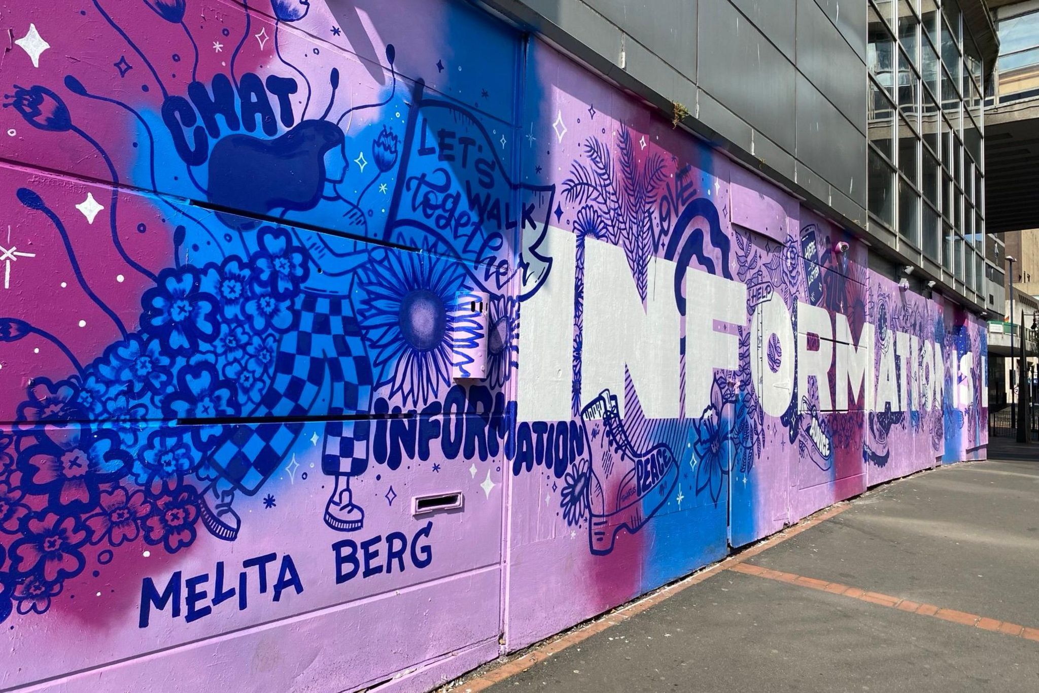 A picture of the mural at an angle. The mural is lilac, dark purple, and dark blue in colour, with 'information is' seen painted on in white lettering as well as 'let's walk together'