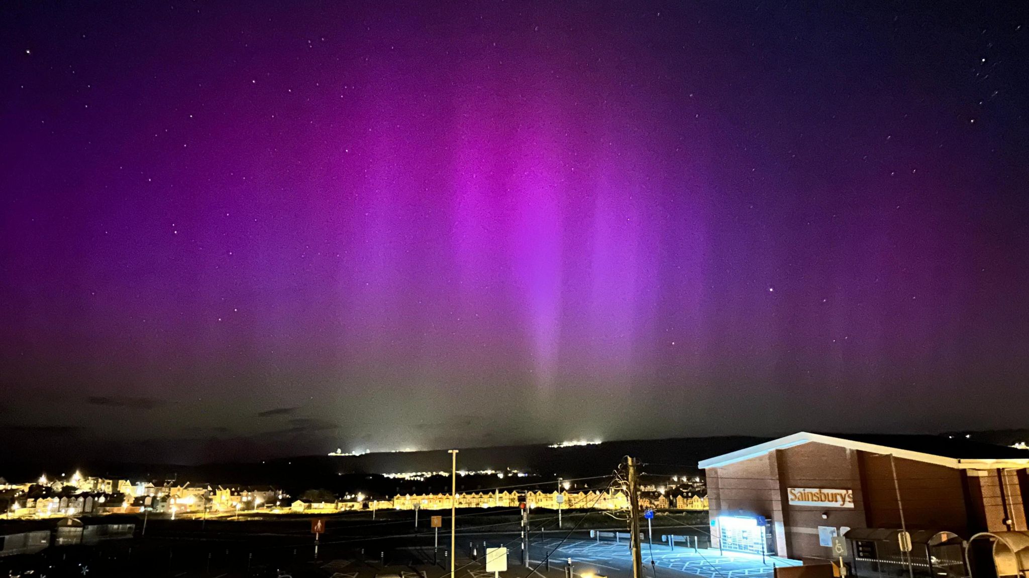 the night sky with buildings and street lights at the bottom of the image with deep and light purple colours of the aurora filling the sky above.