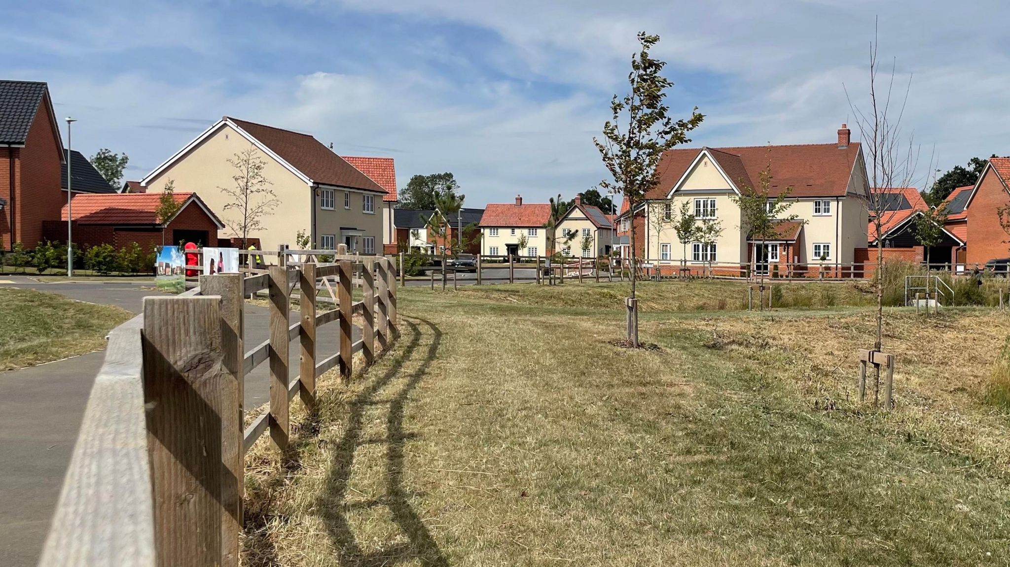 New detached homes in the Beacon Green estate. The homes mostly have cream walls and red tiled roofs, but a few are red brick, with grey tiled roofs. A wooden fence leads the eye into the image, and there is grass and young trees in the foreground.